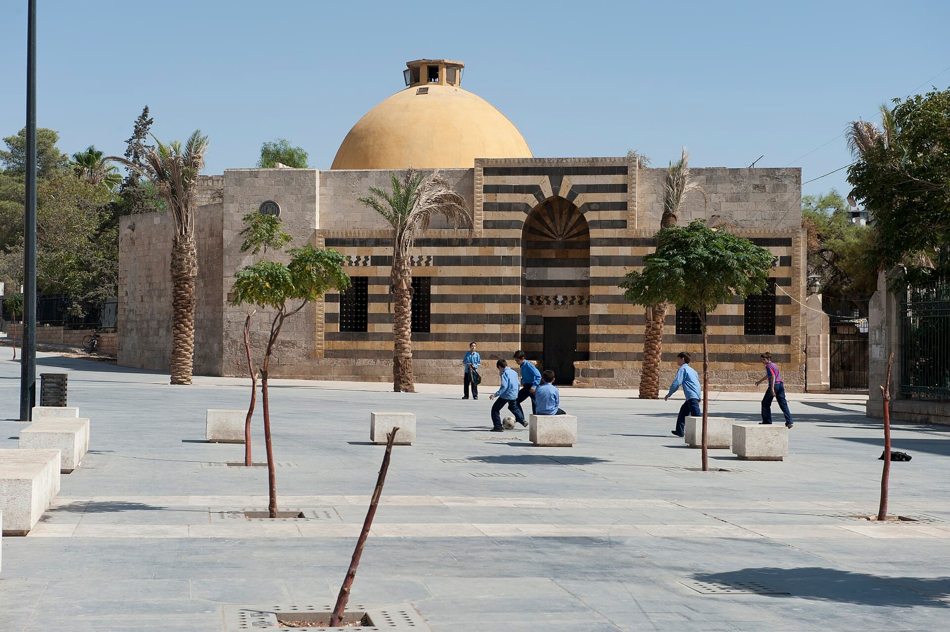 Hamam Yalbuga'nın siyah-sarı taş şeritli cephesi ve sarı kubbesi; önünde kale meydanı