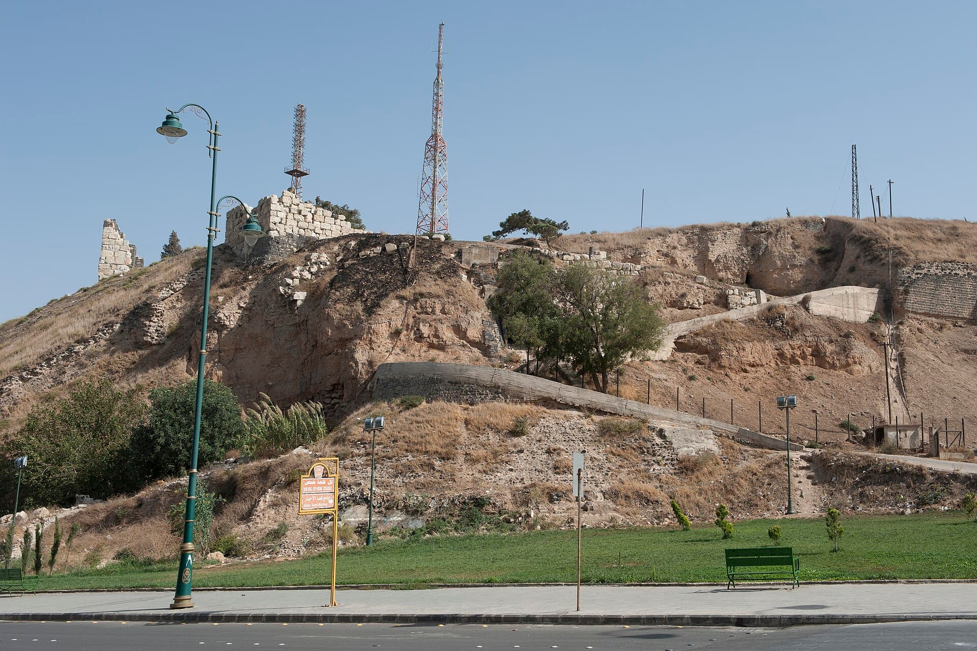 Humus Kalesi höyüğü: taş kaplı yamaç, tepede sur kalıntıları ve anten direkleri, aşağıda bulvar ve park