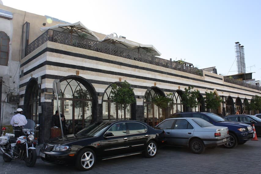 Naranj Lokantası'nın sokak cephesi: siyah-beyaz sıralı ablak taş duvar, alt katta kemerli camekânlar, üst katta şemsiyeli teras, Şam