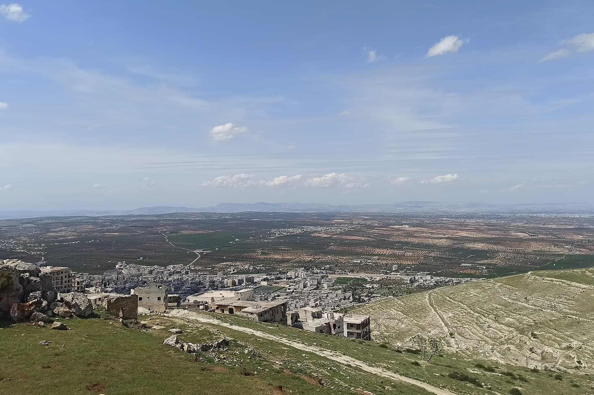 Erbain Dağı'nın yamacından Ariha'ya bakış: önde taşlık patika ve yamaç evleri, aşağıda kasaba ve zeytinliklerle kaplı İdlib ovası