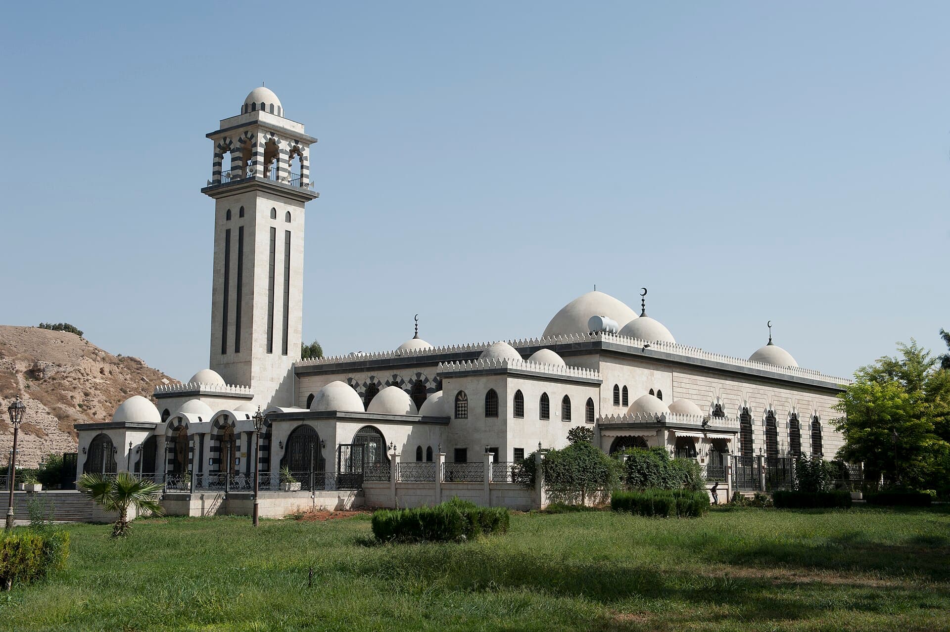Atasi Camii'nin beyaz taş cephesi, kubbe dizisi ve kare minaresi; solda Humus Kalesi'nin höyüğü