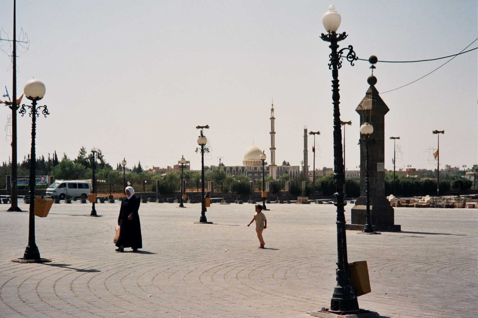 Busra'da taş döşeli geniş bir meydan: dökme demir sokak lambaları, yürüyen bir kadın ve çocuk, arkada kubbeli ve minareli bir cami, 2005