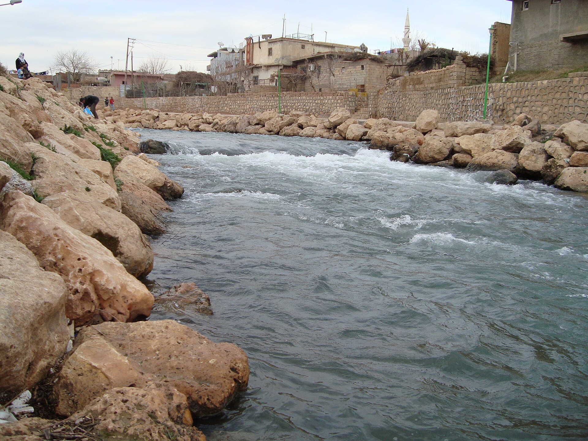 Nusaybin'de, sınırın Türkiye yakasında taş dolgulu kıyılar arasında hızlı akan Cağcağ Nehri; arkada evler ve bir minare