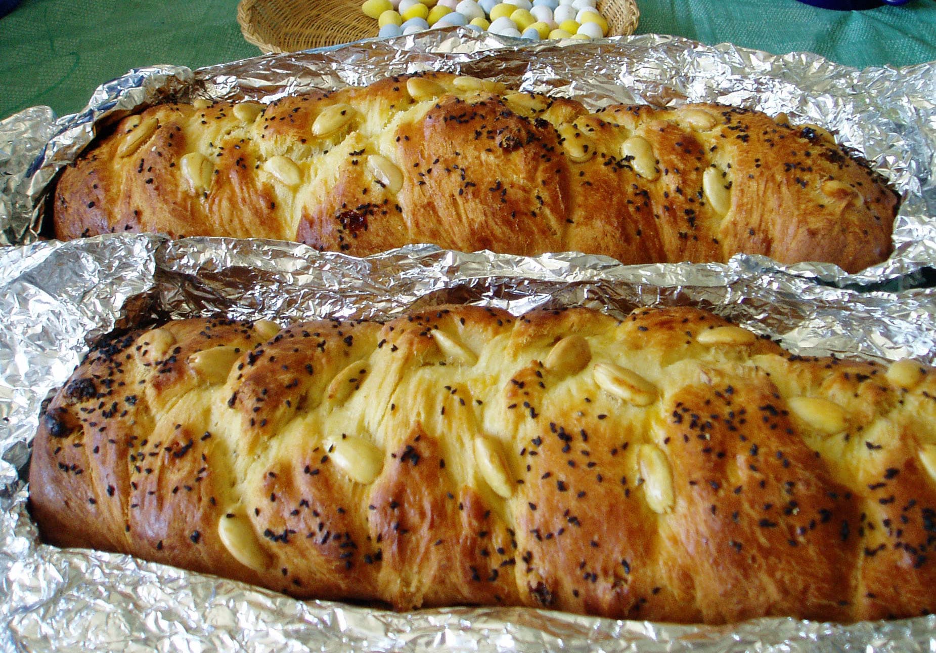 Susam ve çörek otu serpilmiş iki örgülü çörek somunu; arkada Paskalya sepeti duruyor