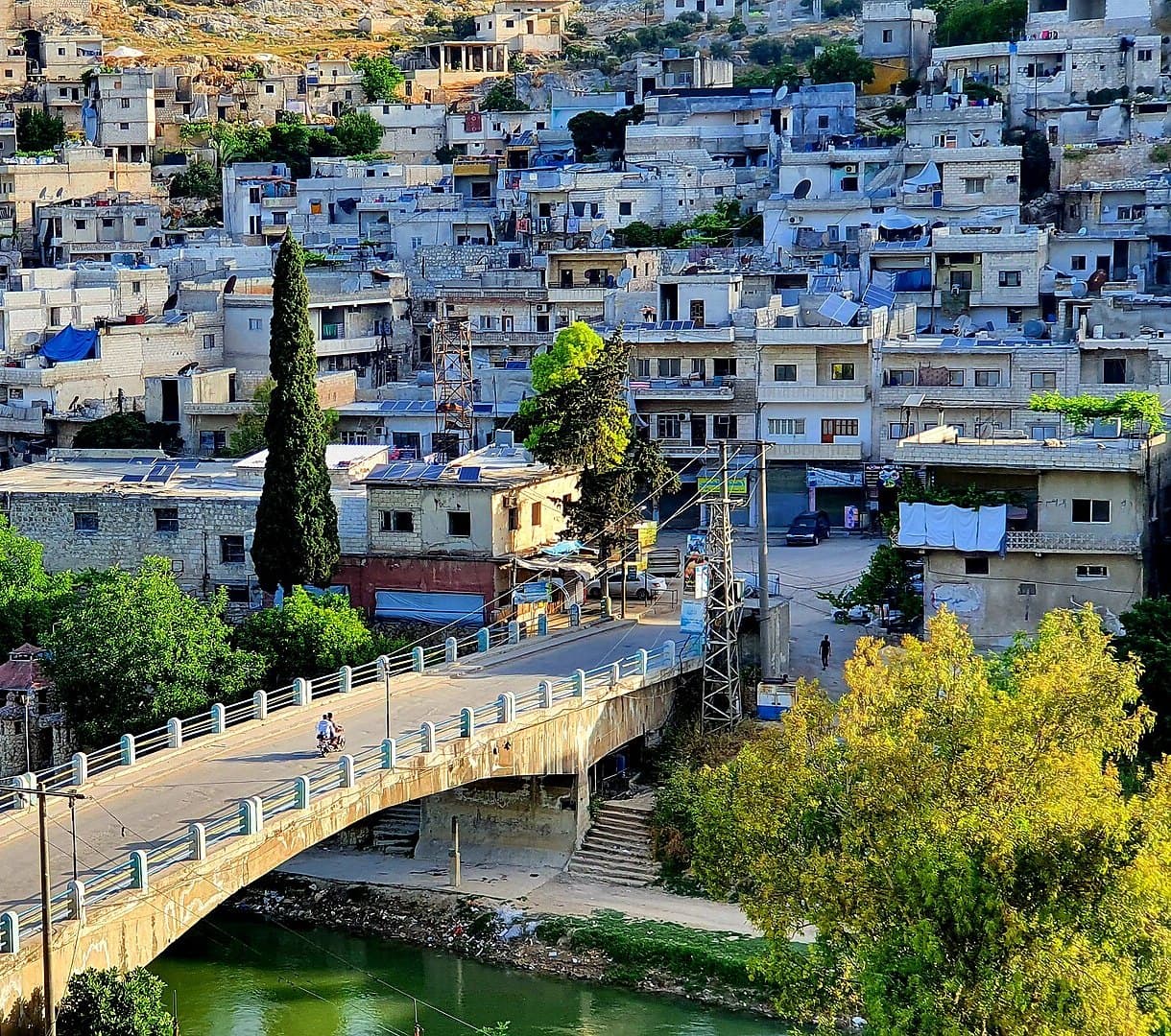 Darkuş'ta Asi Nehri üstündeki modern beton köprü ve arkasında yamaca tırmanan beyaz taş evler, ön planda selvi ve yeşil ağaçlar
