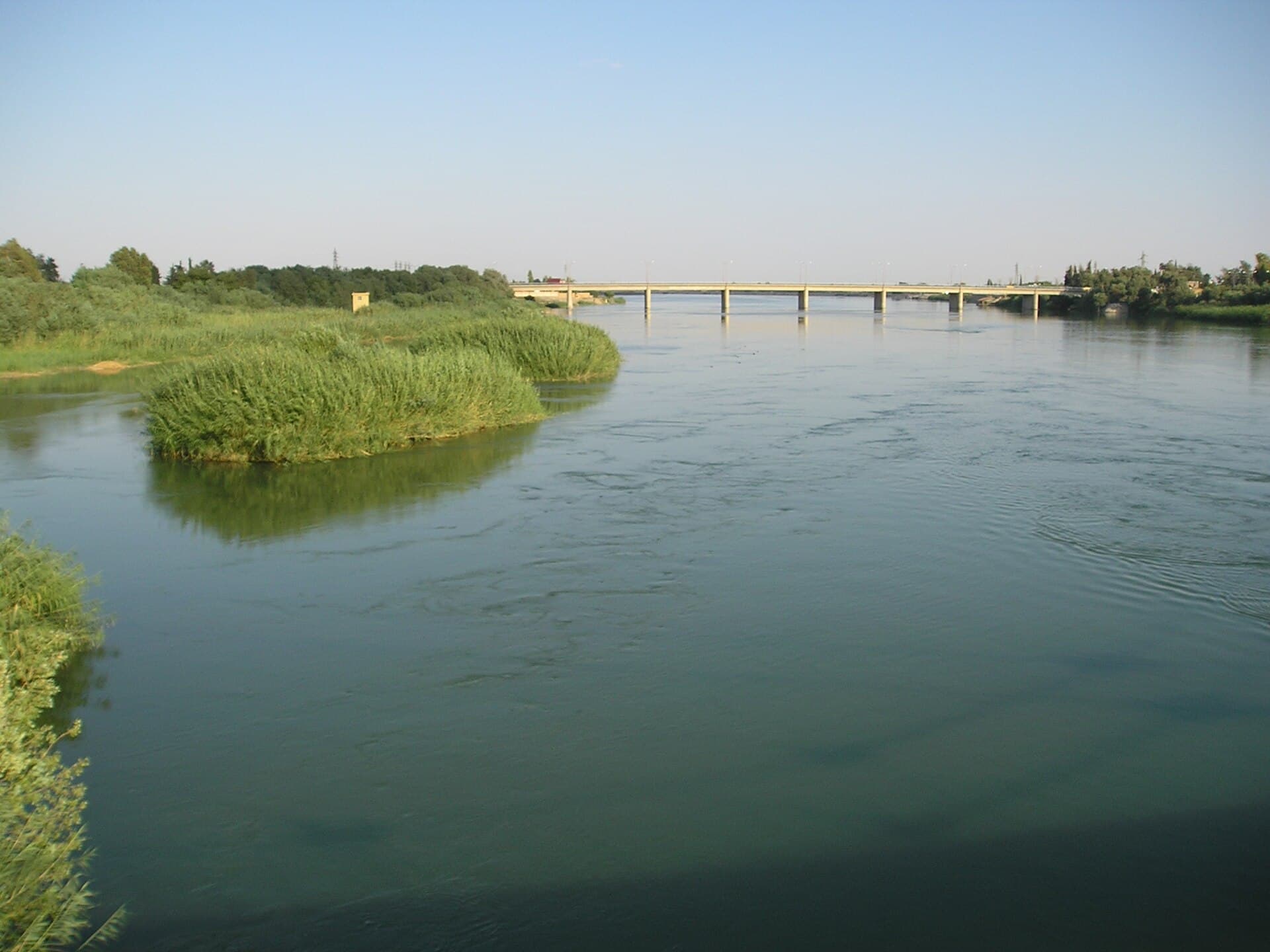 Deyrizor'da Fırat, 2005 yazı: geniş ve sakin nehirde sazlık adacıklar, uzakta nehri geçen beton bir karayolu köprüsü