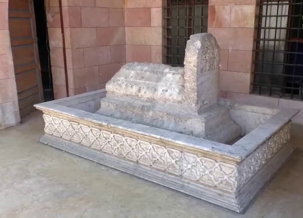Maarat en-Numan'da Ebu'l-Ala el-Maarri'nin mezarı: geometrik desenli mermer çerçeve içinde taş sanduka ve başucunda dikili taş, arkada pembe taş duvar ve demir parmaklıklı pencere