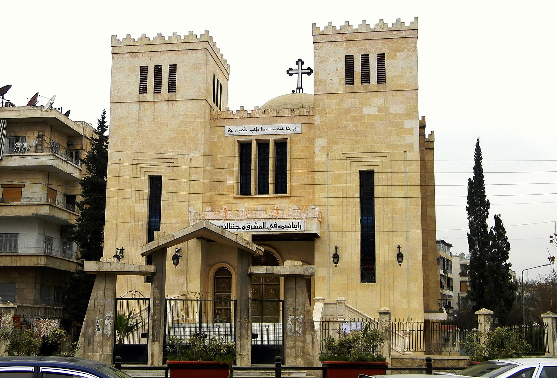 Halep Aziz Yusuf Keldani Katedrali'nin iki mazgallı kule arasındaki cephesi, üstte Süryanice yazı kuşağı ve arkada kubbe
