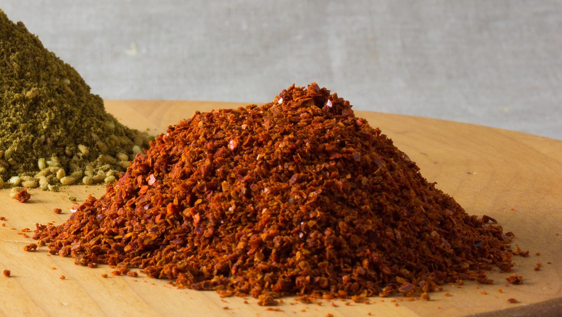 Tahta tezgâhta iri taneli, koyu bordo Halep biberi yığını; arkada zaatar tepeciği