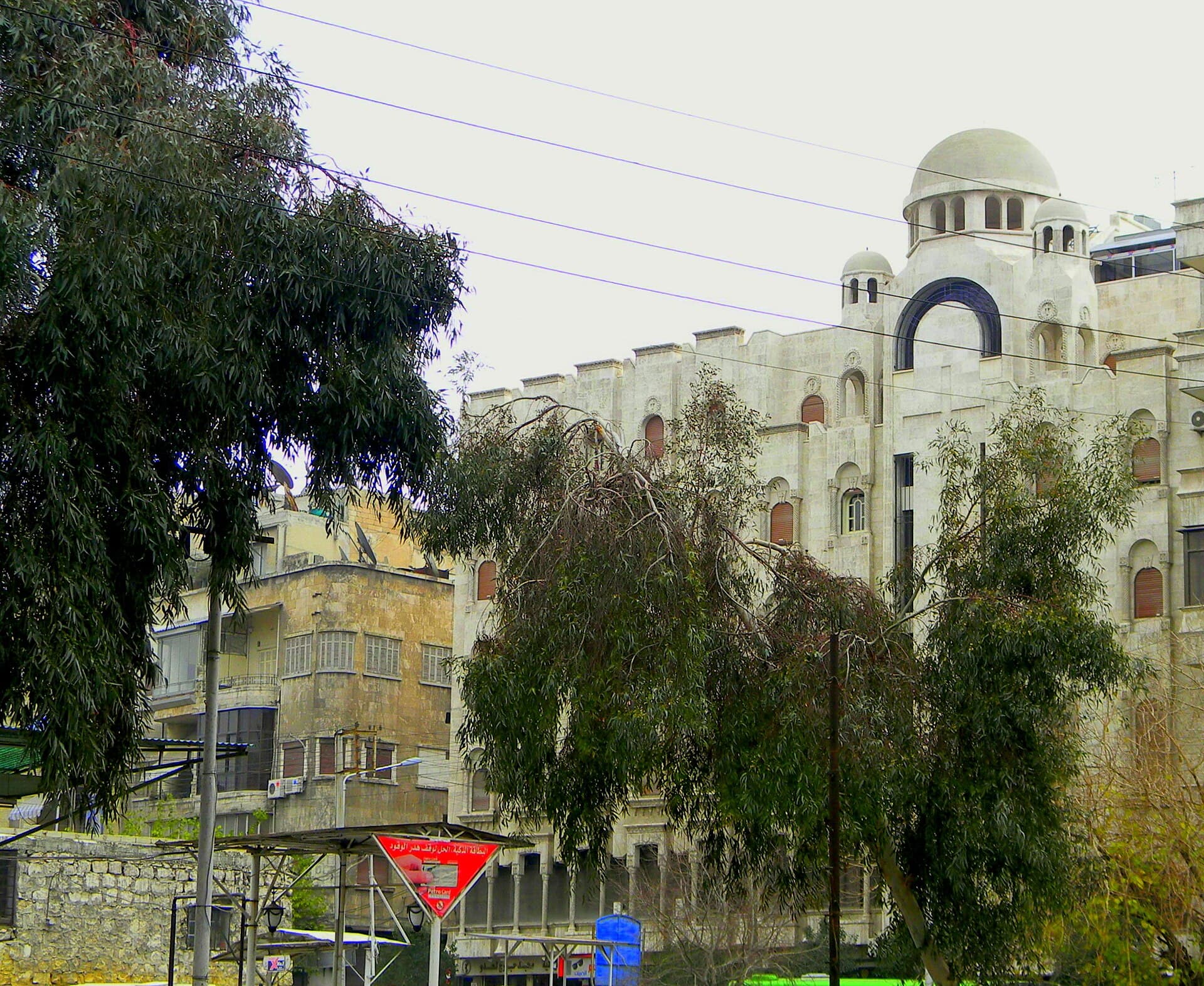 Halep Mar Efrem Katedrali'nin Süleymaniye'deki beyaz taş kütlesi; köşedeki kubbe, kemerli büyük pencere ve önündeki okaliptüsler