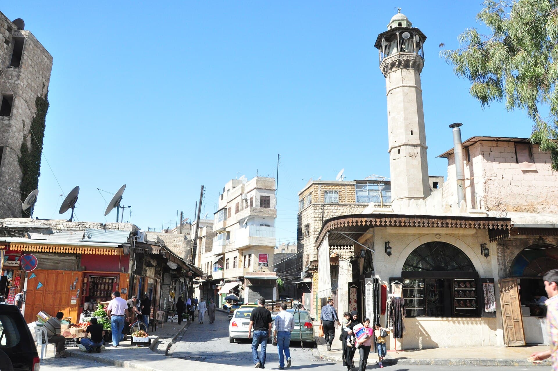 Cdeyde'de Şeref Camii'nin ince taş minaresi; altında ahşap saçaklı çarşı sokağı ve dükkânlar