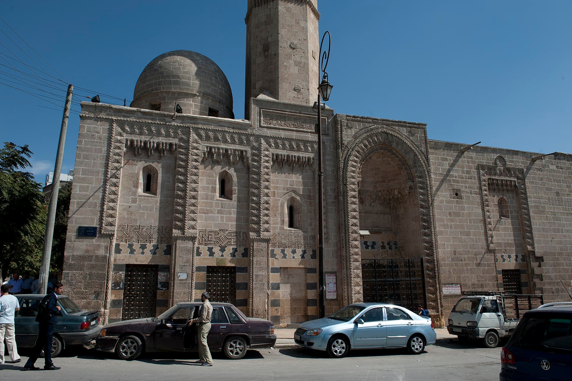 Halep'te Otruş Camii'nin oymalı taş cephesi, sivri kemerli taçkapısı, kubbesi ve minare gövdesi; önünde park etmiş arabalar