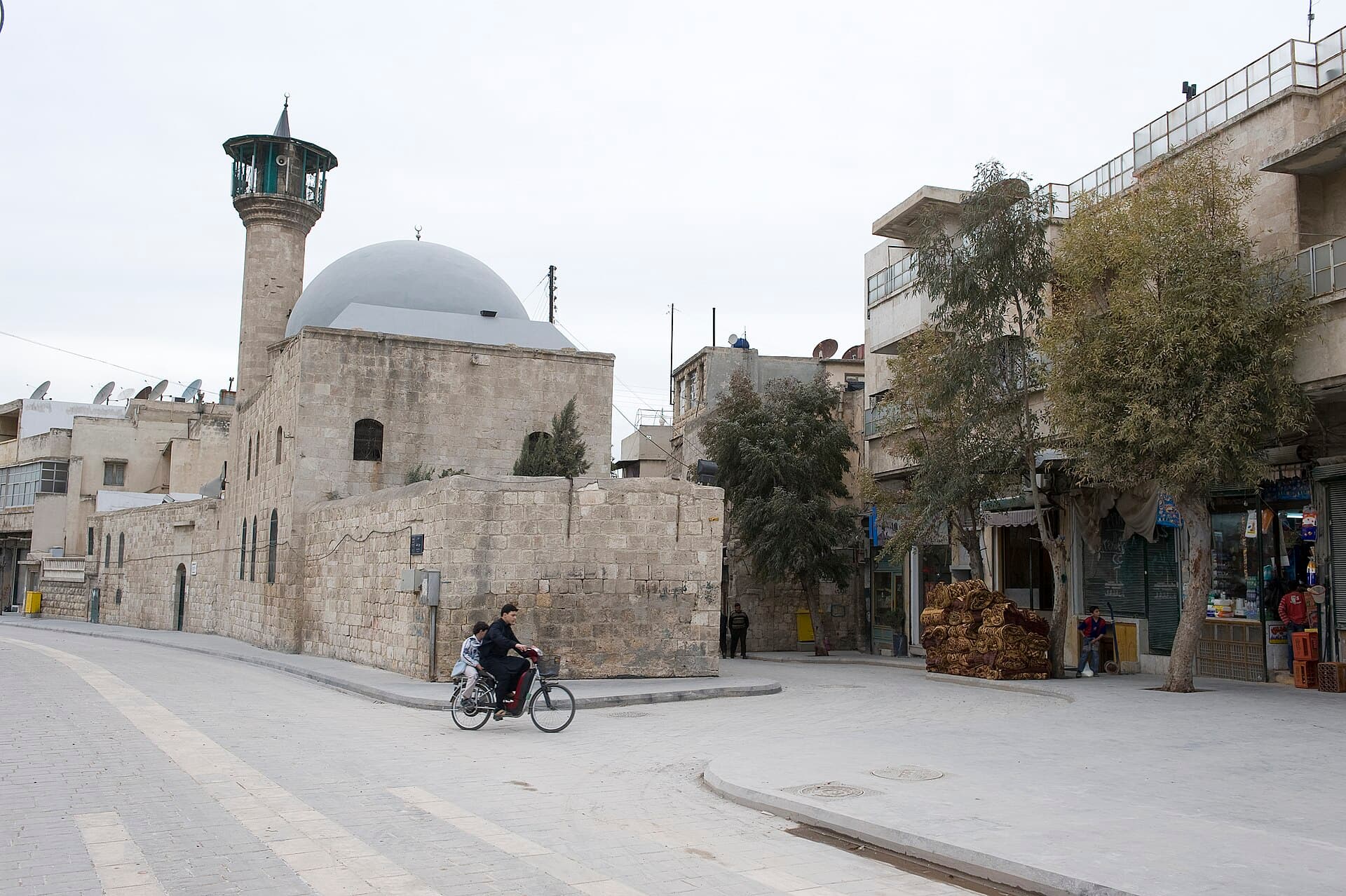 Halep'in Bayâda mahallesinde Hamevi Camii: kesme taş duvarlar, sıvalı kubbe ve kafesli şerefeli minare; önündeki geniş taş sokakta bisikletle geçen iki kişi