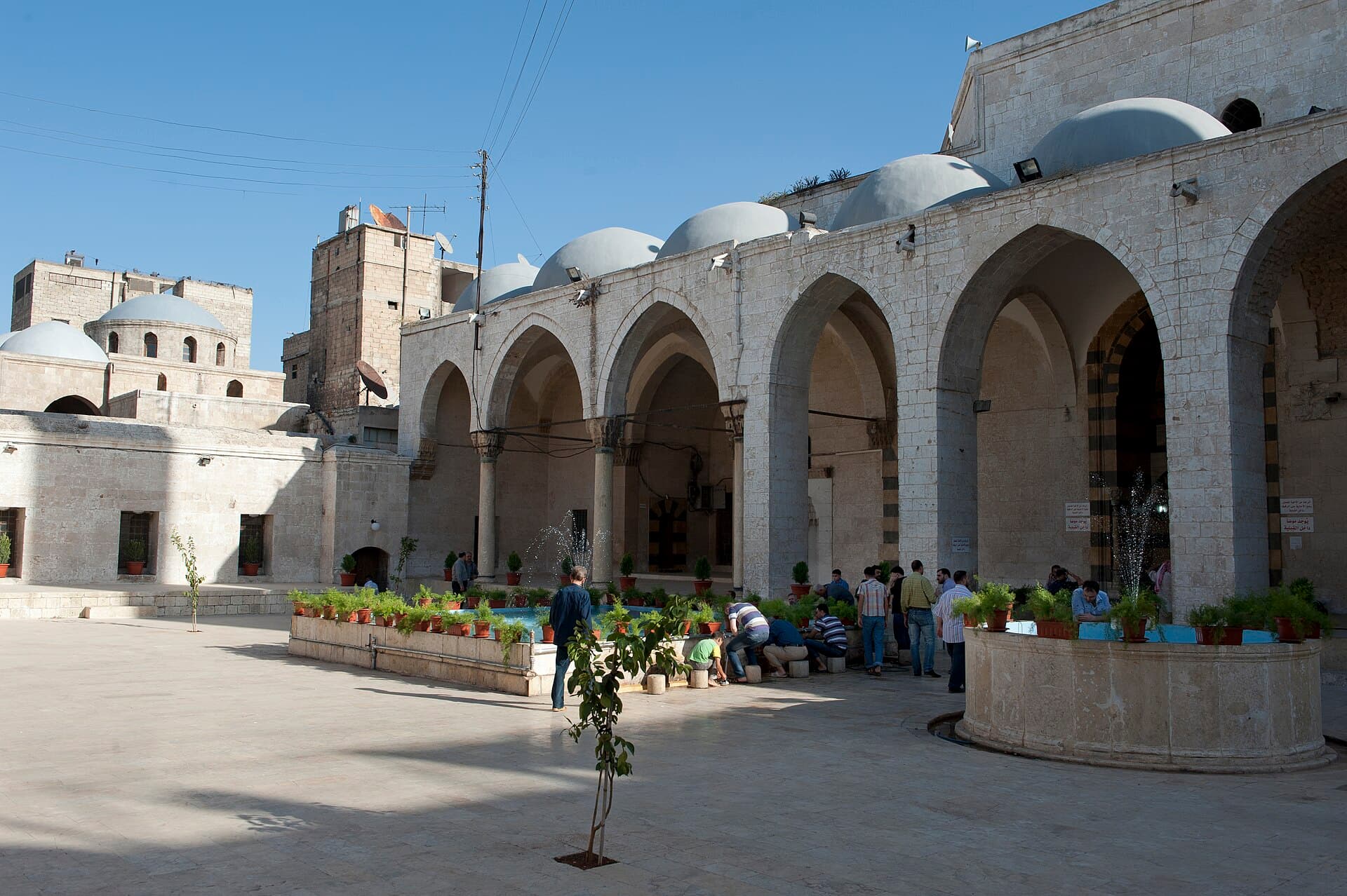Halep'in Cellum mahallesindeki Behramiye Camii'nin avlusu: küçük kubbeli revaklar, saksılarla çevrili şadırvan havuzu ve abdest alan cemaat