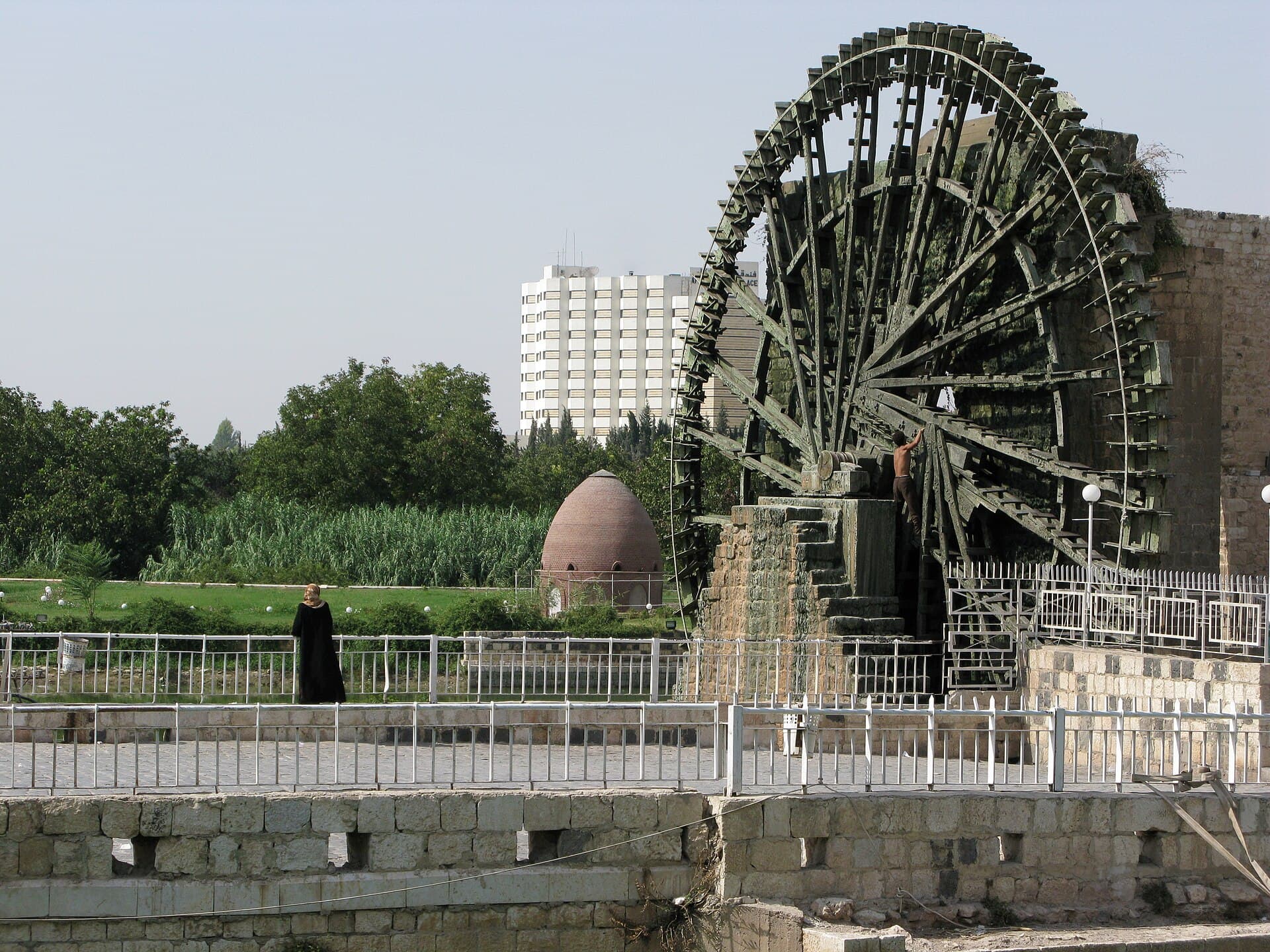 Hama'da Asi Nehri kıyısında taş kaide üstünde duran büyük ahşap su dolabı, önünde korkuluklu kıyı yolu ve arkada kubbeli küçük yapı