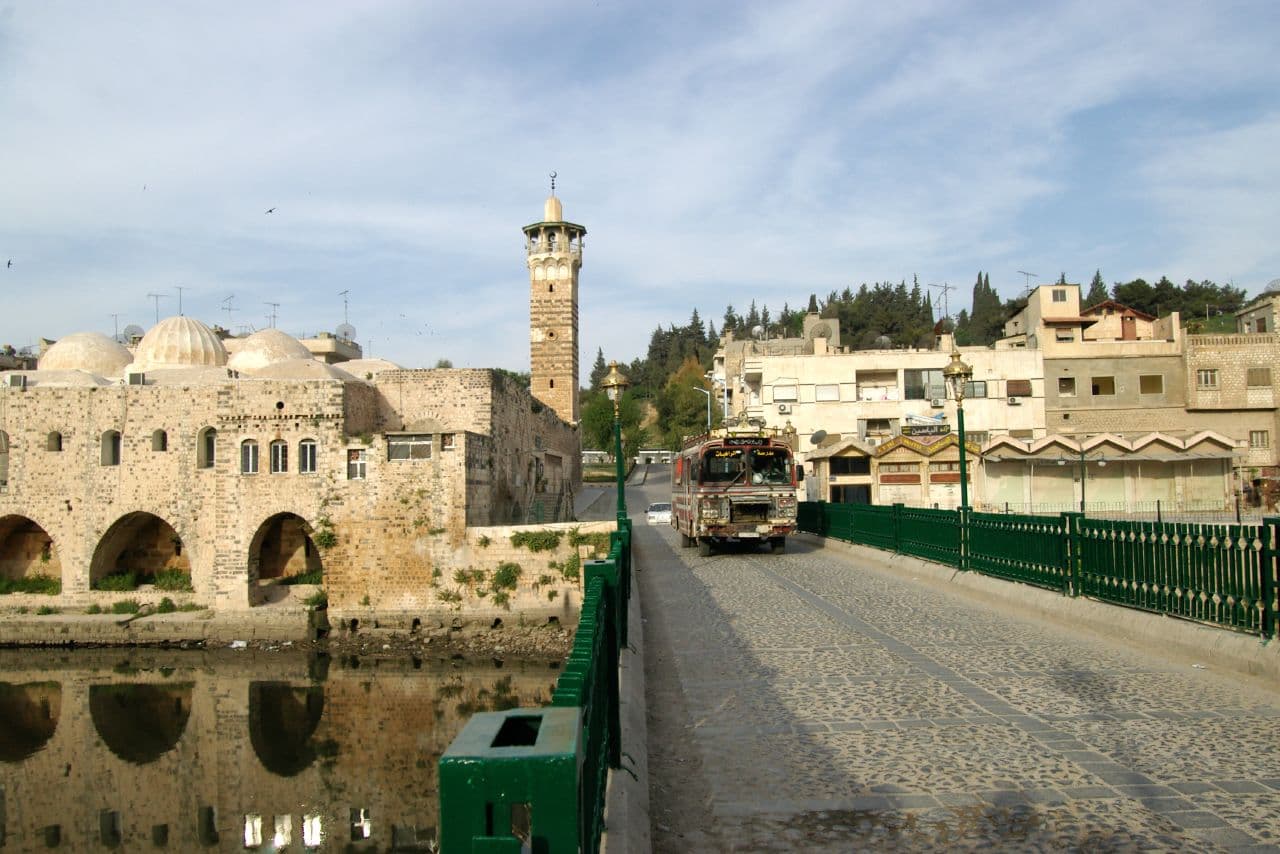 Hama'da Asi Nehri üzerindeki taş döşeli Kilaniyye Köprüsü ve köprünün ucunda kubbeleri ve minaresiyle Nuri Camii