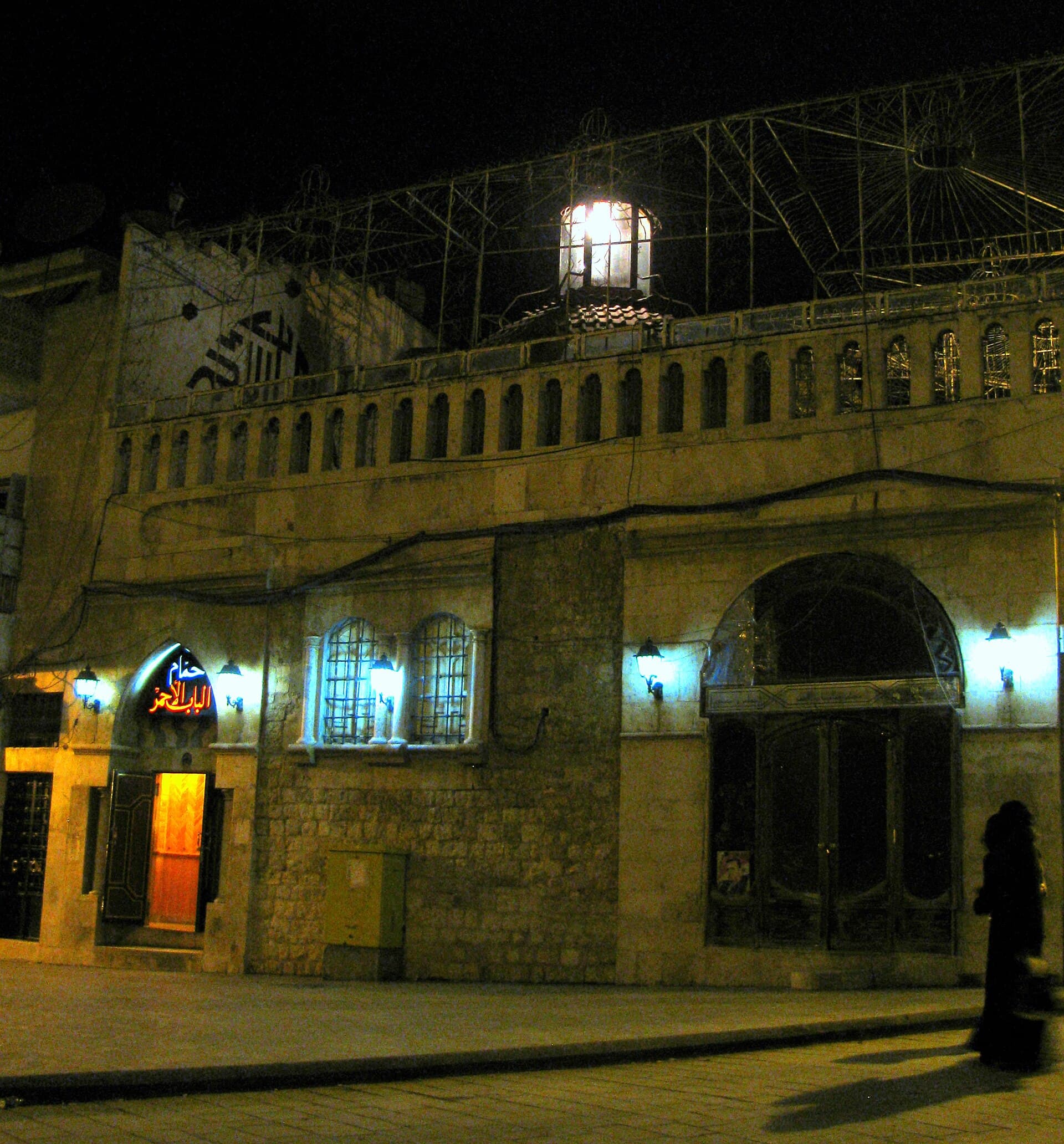 Bab el-Ahmer Hamamı'nın gece aydınlatılmış taş cephesi; kapı üstünde Arapça neon tabela, çatıda fenerli kubbe