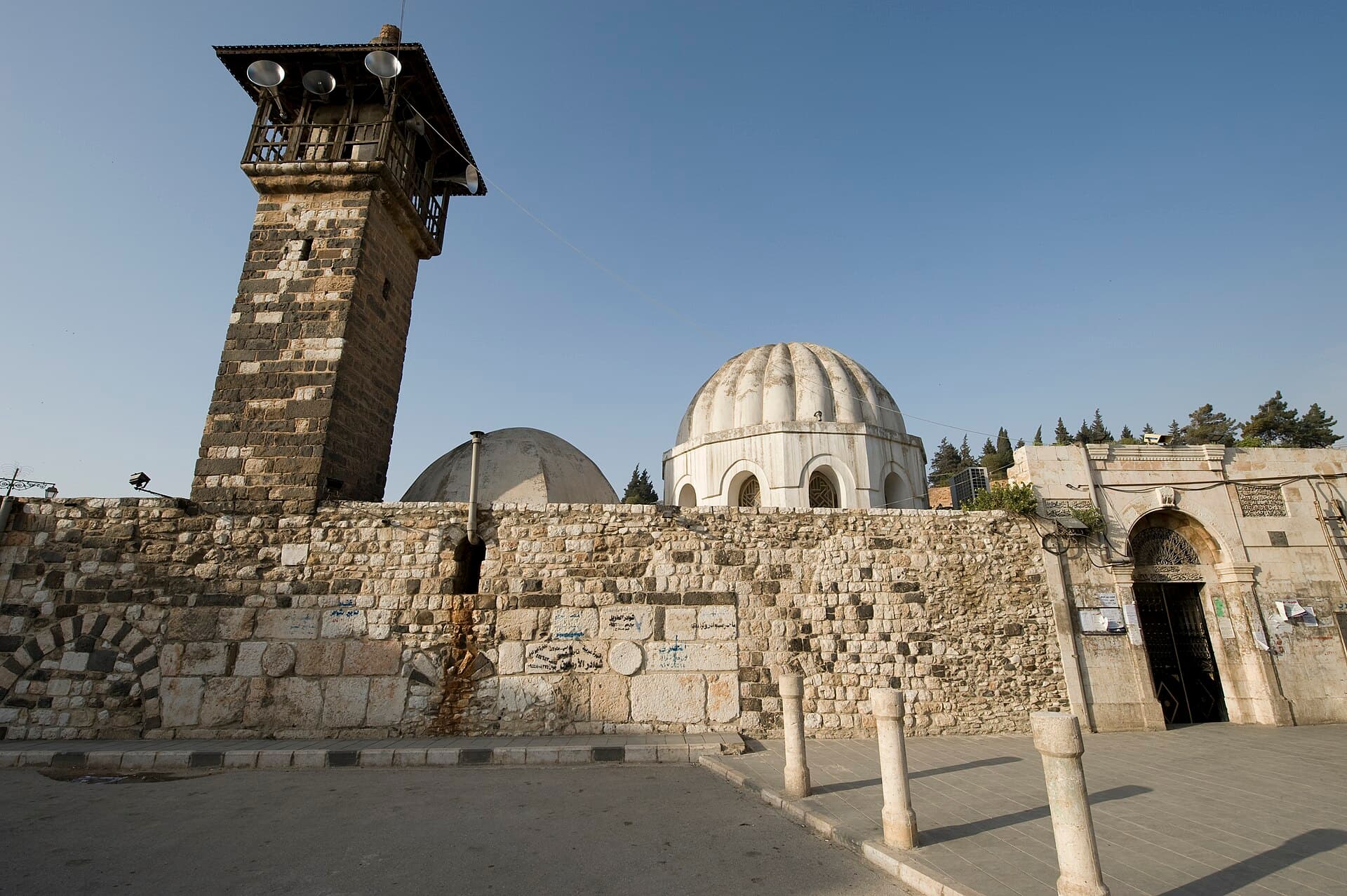 Hama'daki Hasaneyn Camii; siyah beyaz taş sıralı minare, taş duvarın arkasında iki kubbe ve sağda giriş kapısı