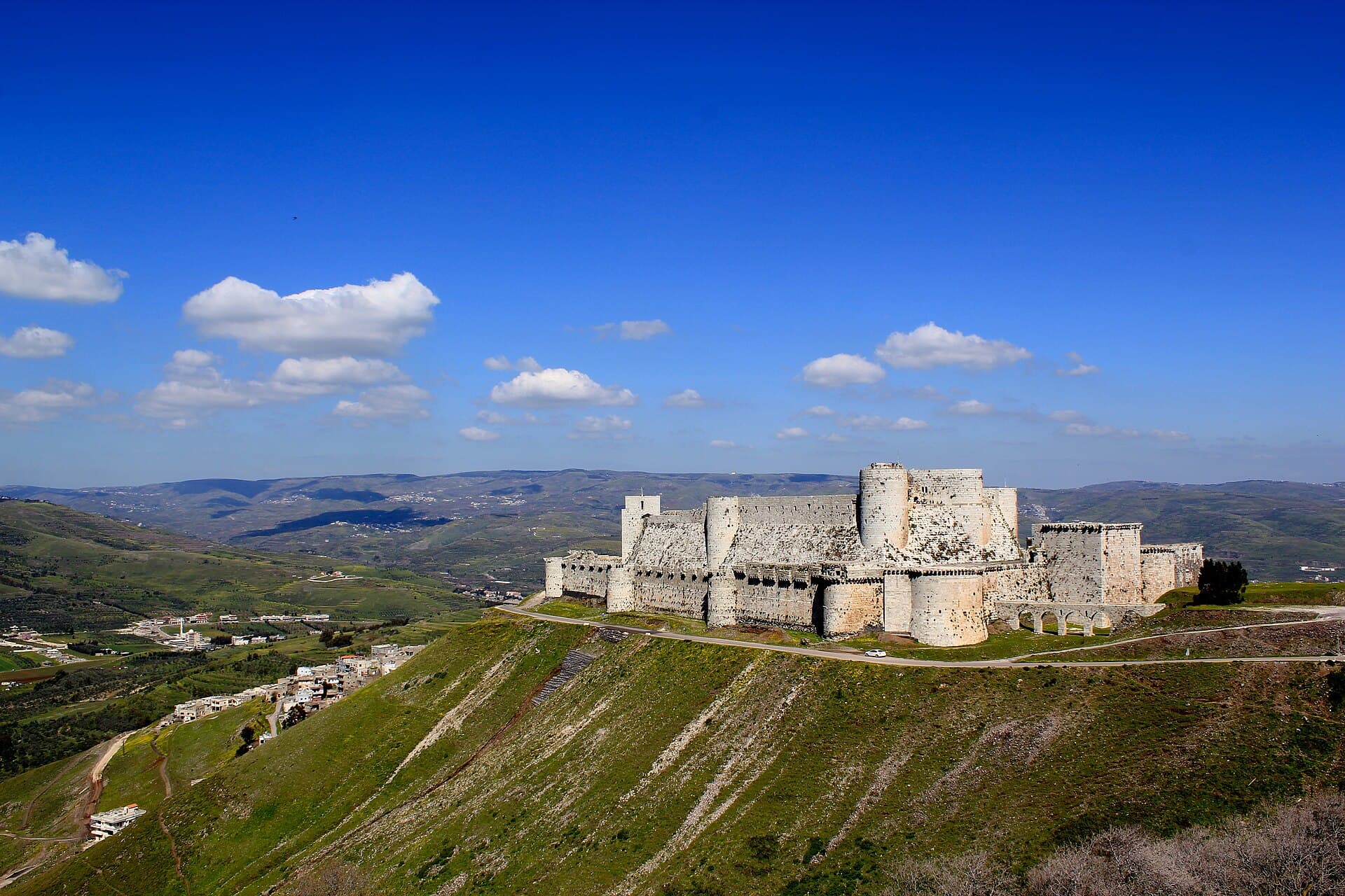 Yeşil sırtın üstünde Krak des Chevaliers ve yamaçtaki köy evleri, arkada vadi ve tepeler
