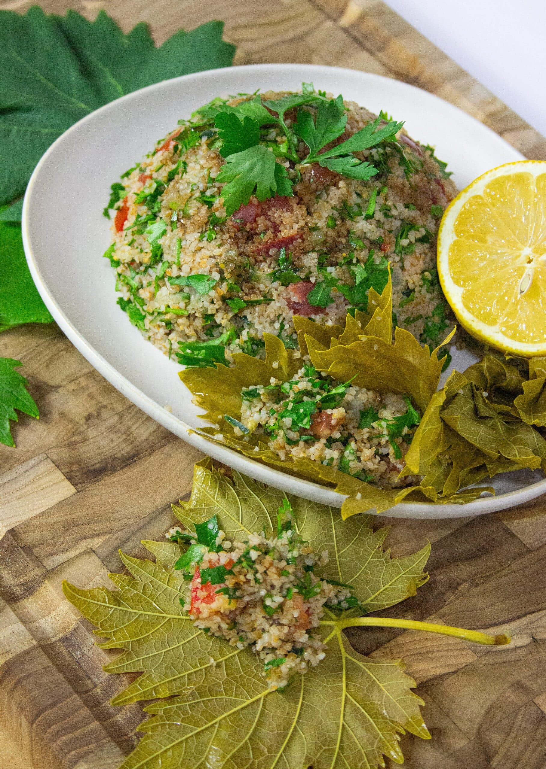 Beyaz tabakta maydanoz ve domatesli bulgur karışımı, yanında kaşıkla doldurulmuş asma yaprakları ve yarım limon