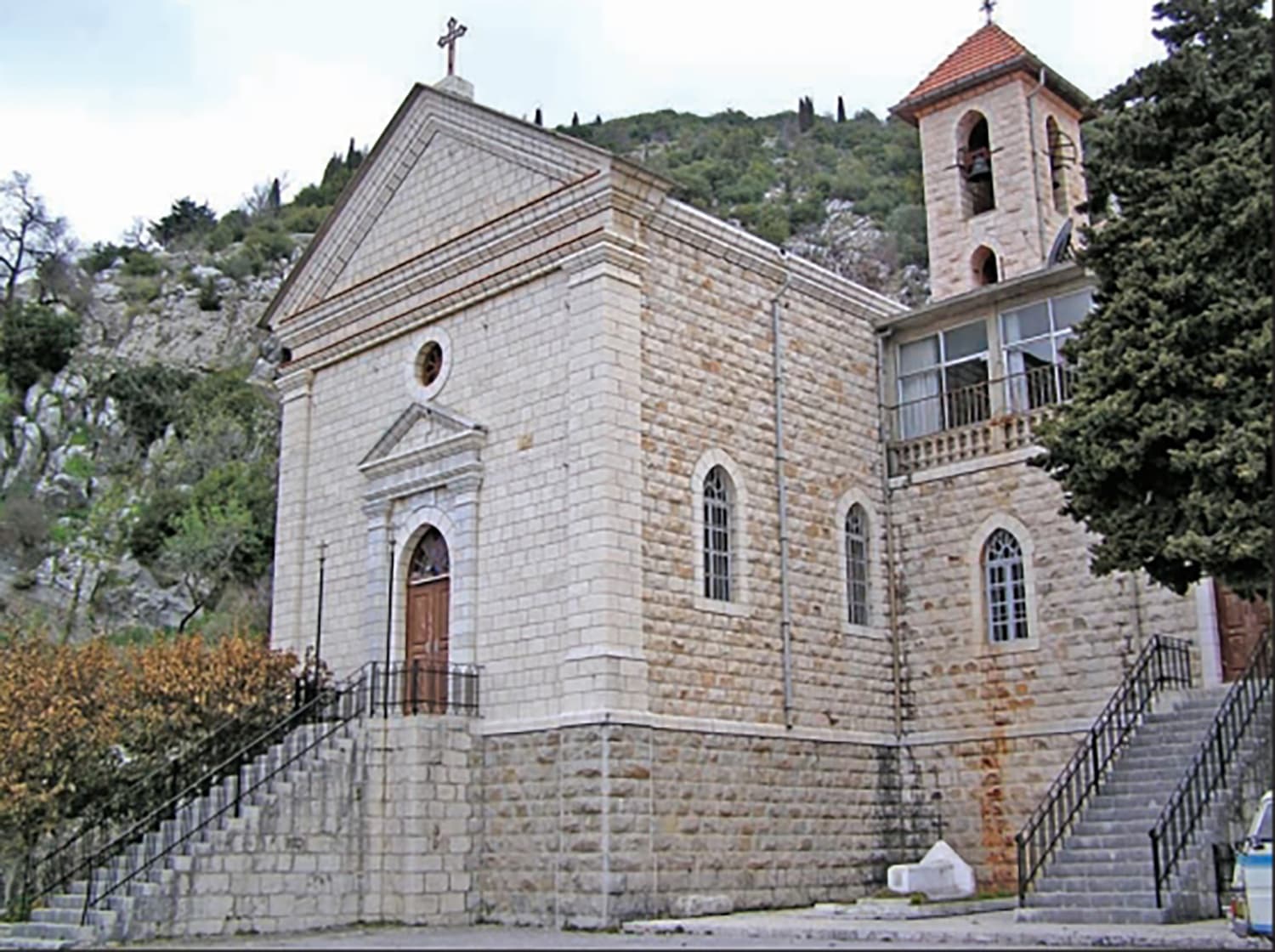 Kessab'daki Aziz Mikail Ermeni Katolik Kilisesi: üçgen alınlıklı kesme taş cephe, merdivenli giriş ve yanındaki çan kulesi, arkada ormanlı kayalık yamaç