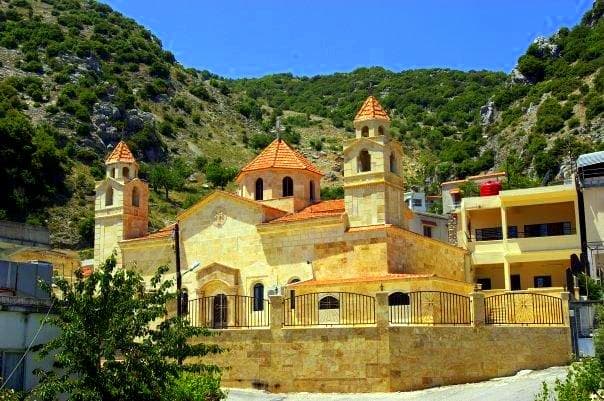 Kessab Meryem Ana Ermeni Kilisesi'nin kubbesi ve iki çan kulesi, arkasında çam ve çalıyla örtülü dağ yamacı