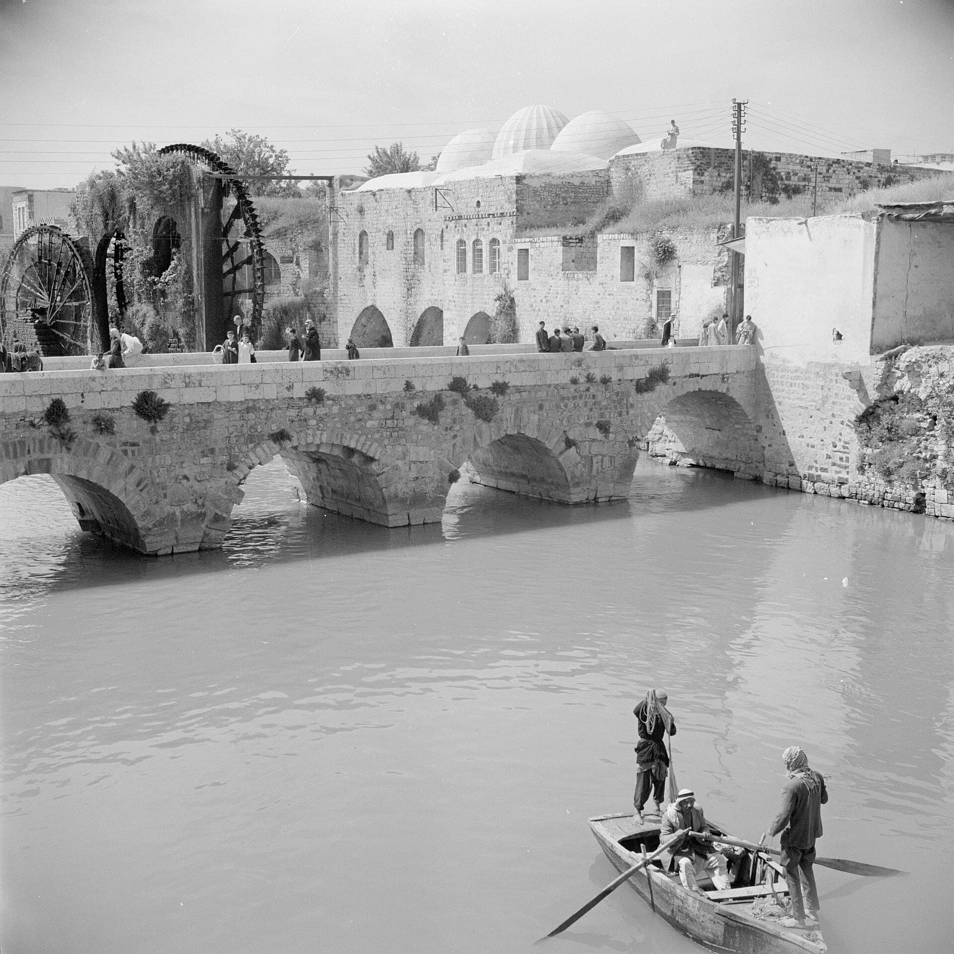 1950'de Hama'da Asi Nehri üzerindeki taş kemerli Kilaniyye Köprüsü; arkada su dolapları ve kubbeli taş yapı, önde nehirde kayıkla geçen üç kişi