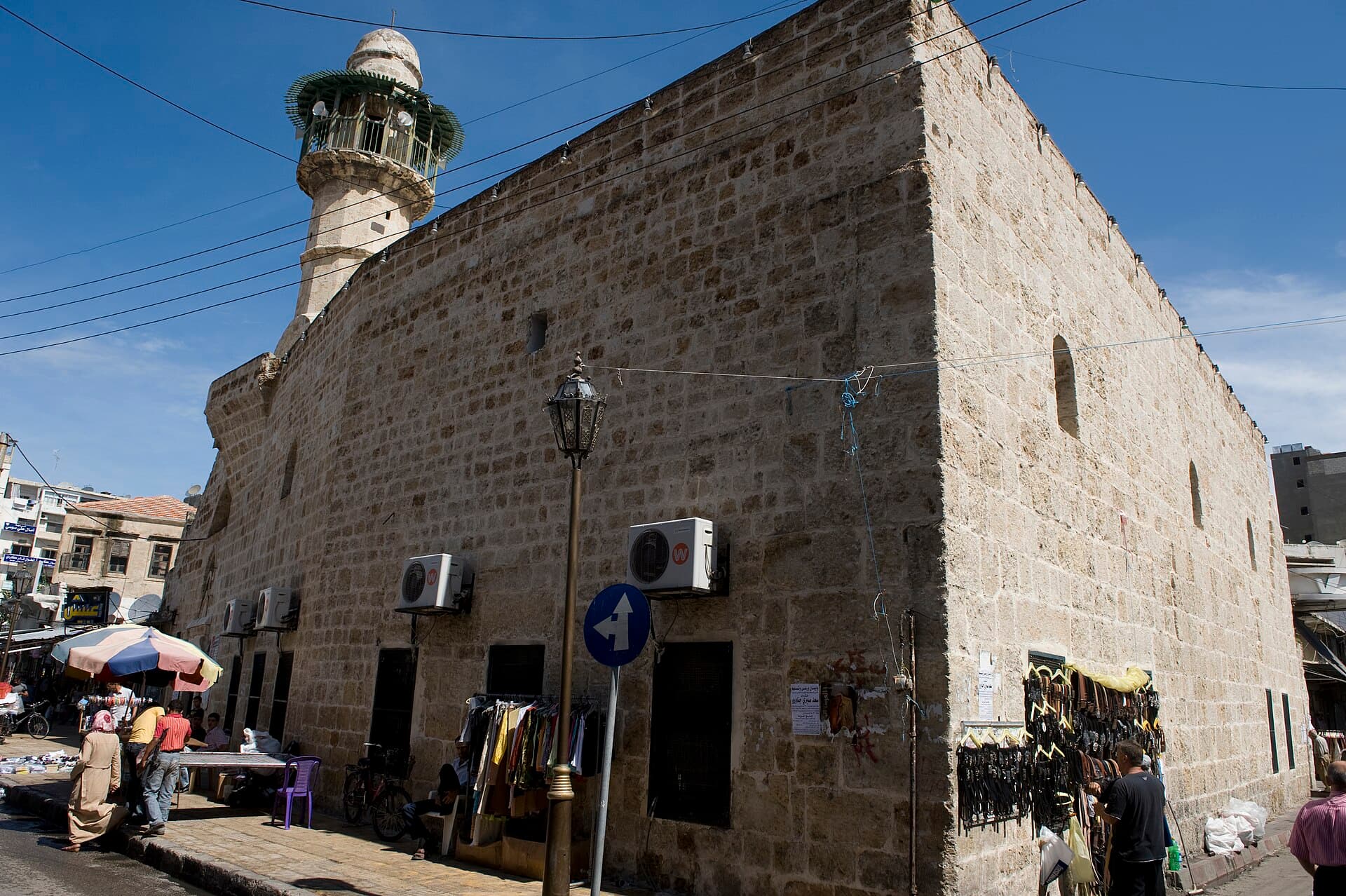 Lazkiye Bazar Camii'nin kesme taş duvarlı dış cephesi ve kafesli şerefeli minaresi; duvar dibinde çarşı tezgâhları ve yoldan geçenler
