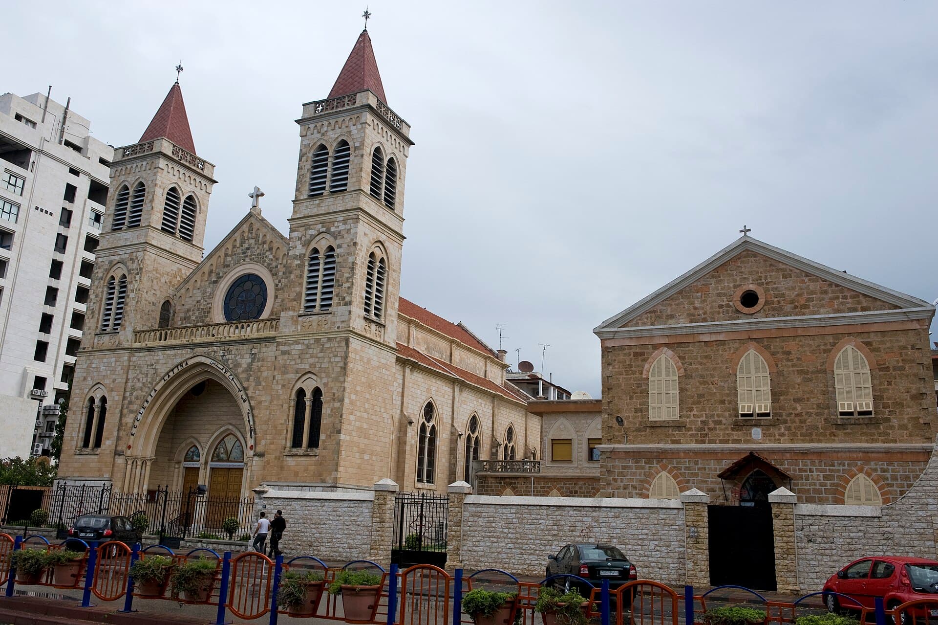 Lazkiye Latin Kilisesi'nin kiremit külahlı iki kulesi ve gül pencereli taş cephesi; sağında aynı parmaklığın arkasında alınlıklı sade bir taş bina