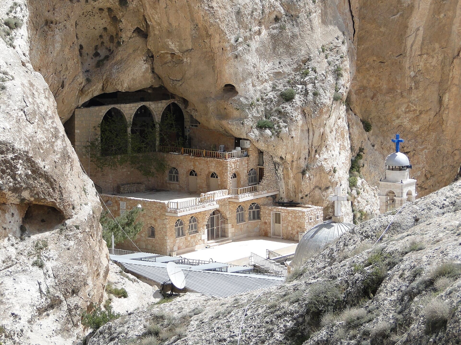 Malula'da Mar Tekla Manastırı: kayanın oyuğuna yerleşmiş kemerli taş bina, önde kubbeli kilise ve mavi haçlı çan kulesi