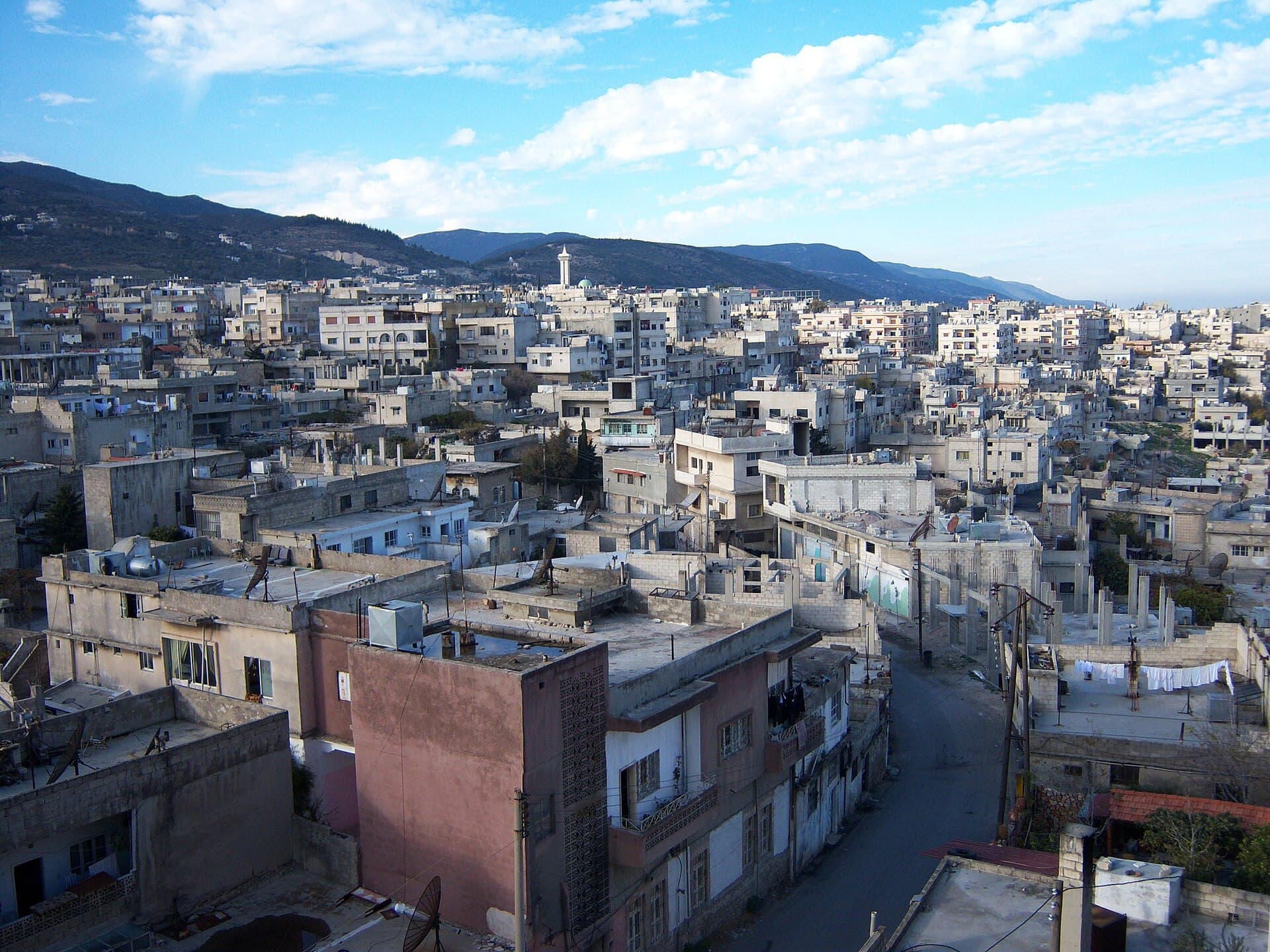 Masyaf kasabasının damları yukarıdan; sıkışık açık renkli beton evler, ortada bir minare ve arkada dağ sırası