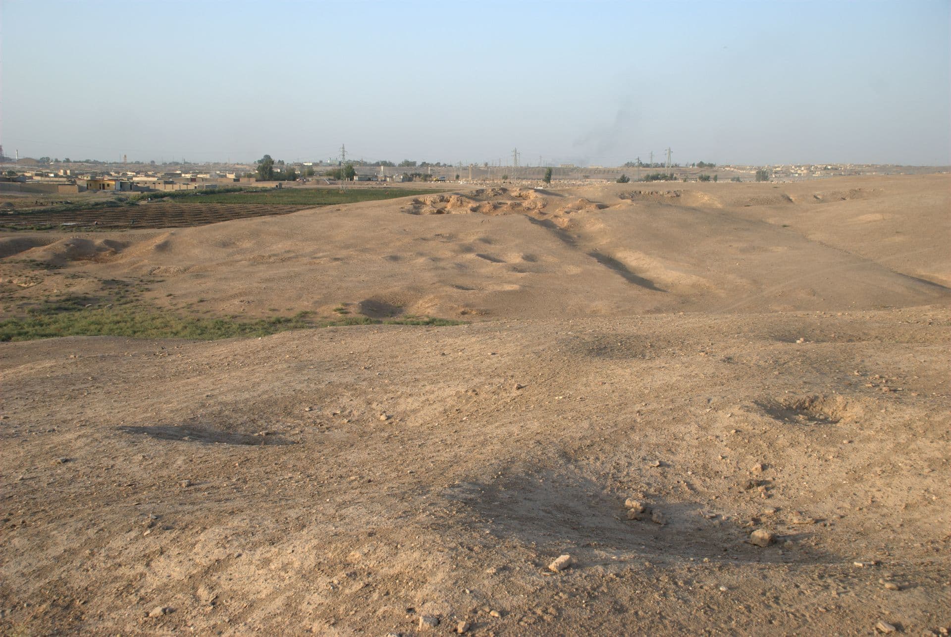 Rakka'nın kuzeydoğusunda Tuttul (Tell Bia) höyüğünün kaçak kazı çukurlu yüzeyi, ufukta kentin evleri ve elektrik direkleri