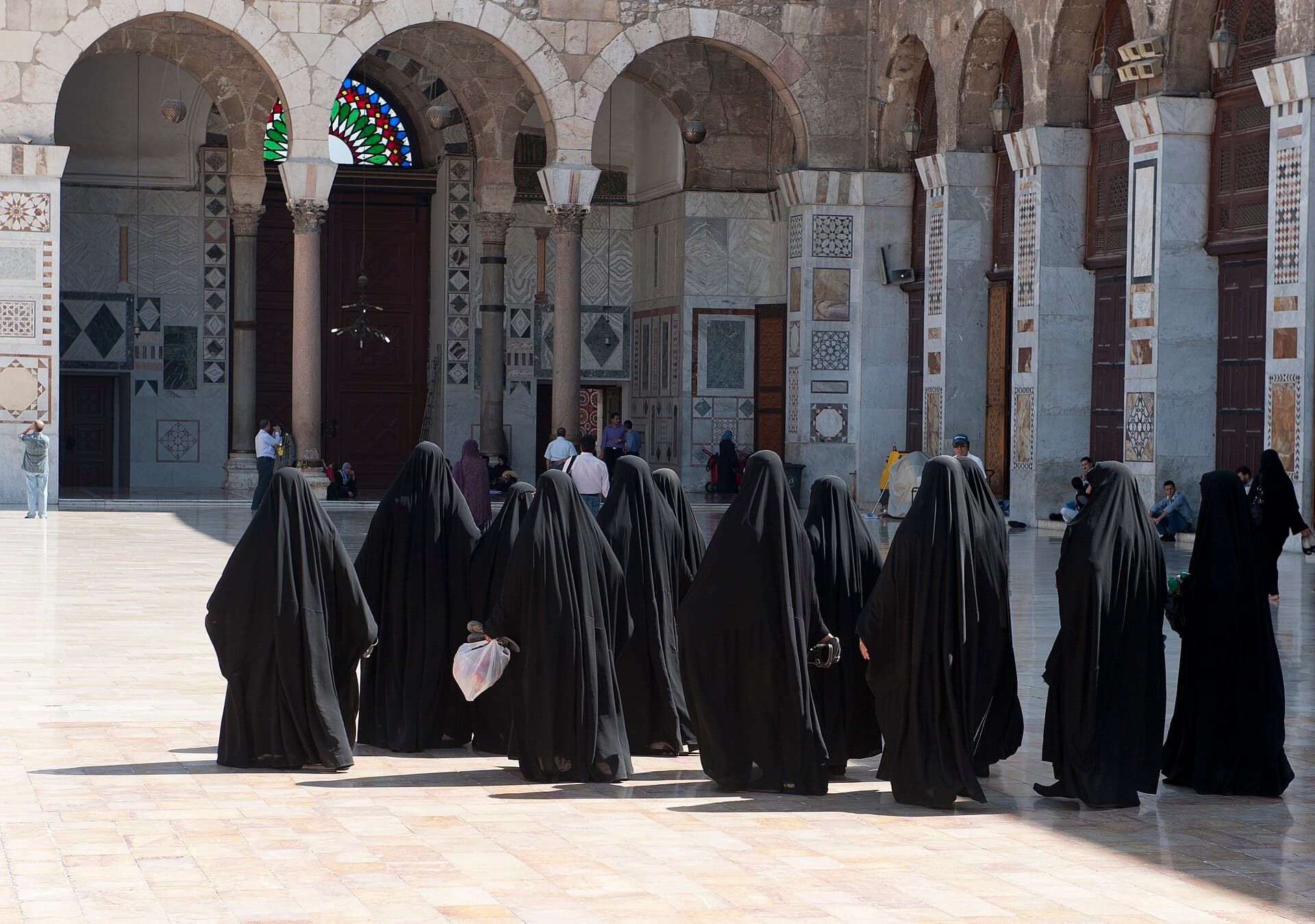 Emevî Camii avlusunda uzun siyah örtüleriyle yürüyen bir ziyaretçi grubu; arkada mermer kaplı revak ve renkli vitray