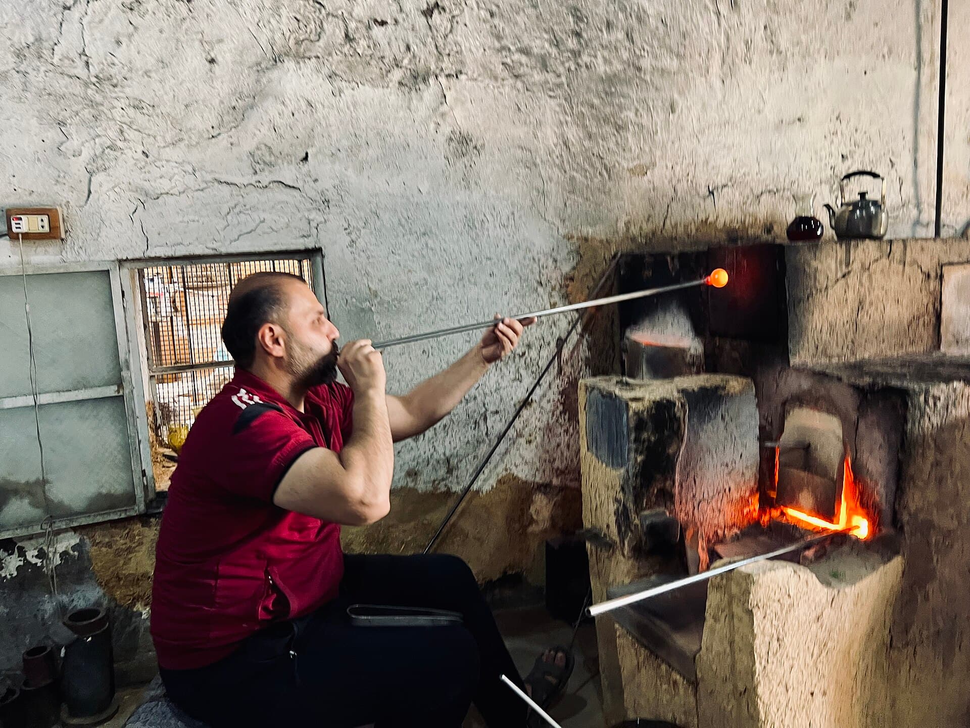 Şam'da bir cam üfleme atölyesi; usta elle örülmüş tuğla fırının önünde oturmuş, demir çubuğun ucundaki kızgın cama üflüyor