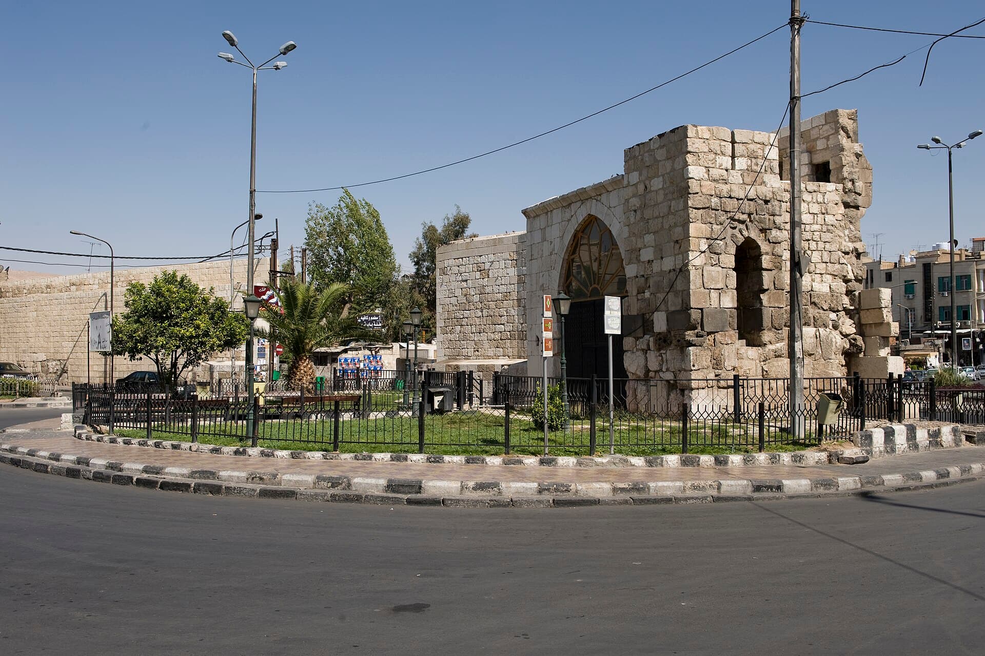 Şam'da Bab Tuma kapısı, surun dışındaki meydandan bakışla: kemerli geçidi ve yıpranmış taş kulesiyle kapı, önünde çitle çevrili yeşil ada ve boş yol