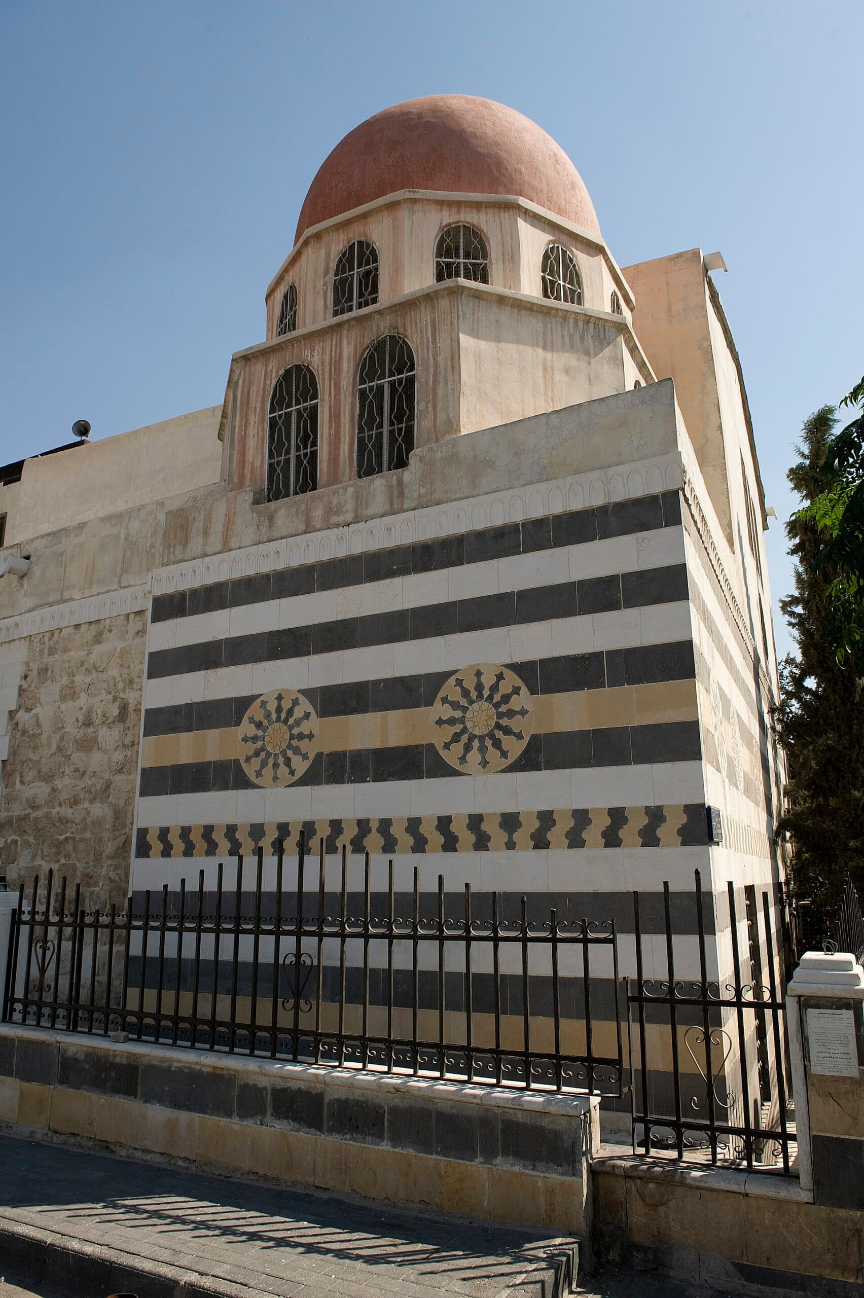 Şam Kanavat'taki Şazbekiyye Camii'nin siyah-beyaz taş bantlı, iki taş rozetli köşe cephesi ve üstünde pencereli kasnağa oturan kırmızı kubbe