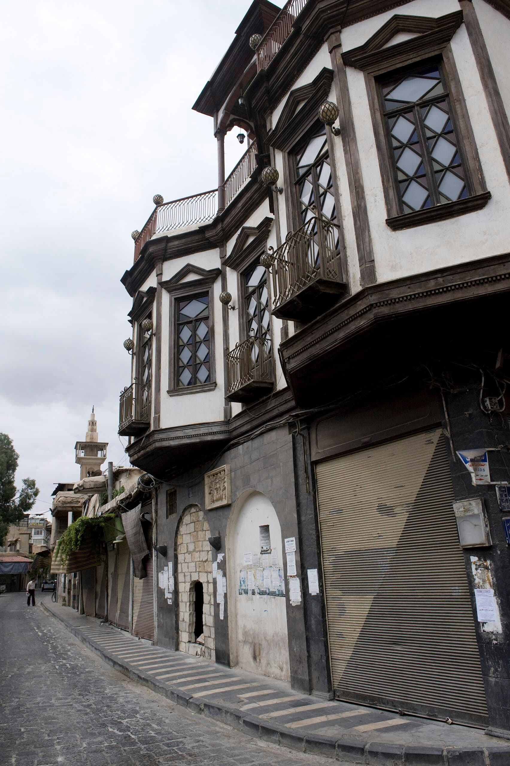 Suk Saruca'da sokağa taşan ahşap üst katlı Osmanlı evi; arka planda minare