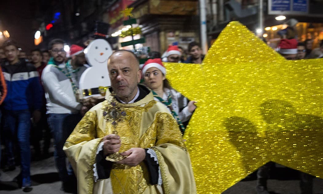 Şam'da Bab Tuma'daki Noel alayında altın işlemeli cüppeli bir rahip, arkasında Noel Baba şapkalı gençler ve büyük parlak bir yıldız