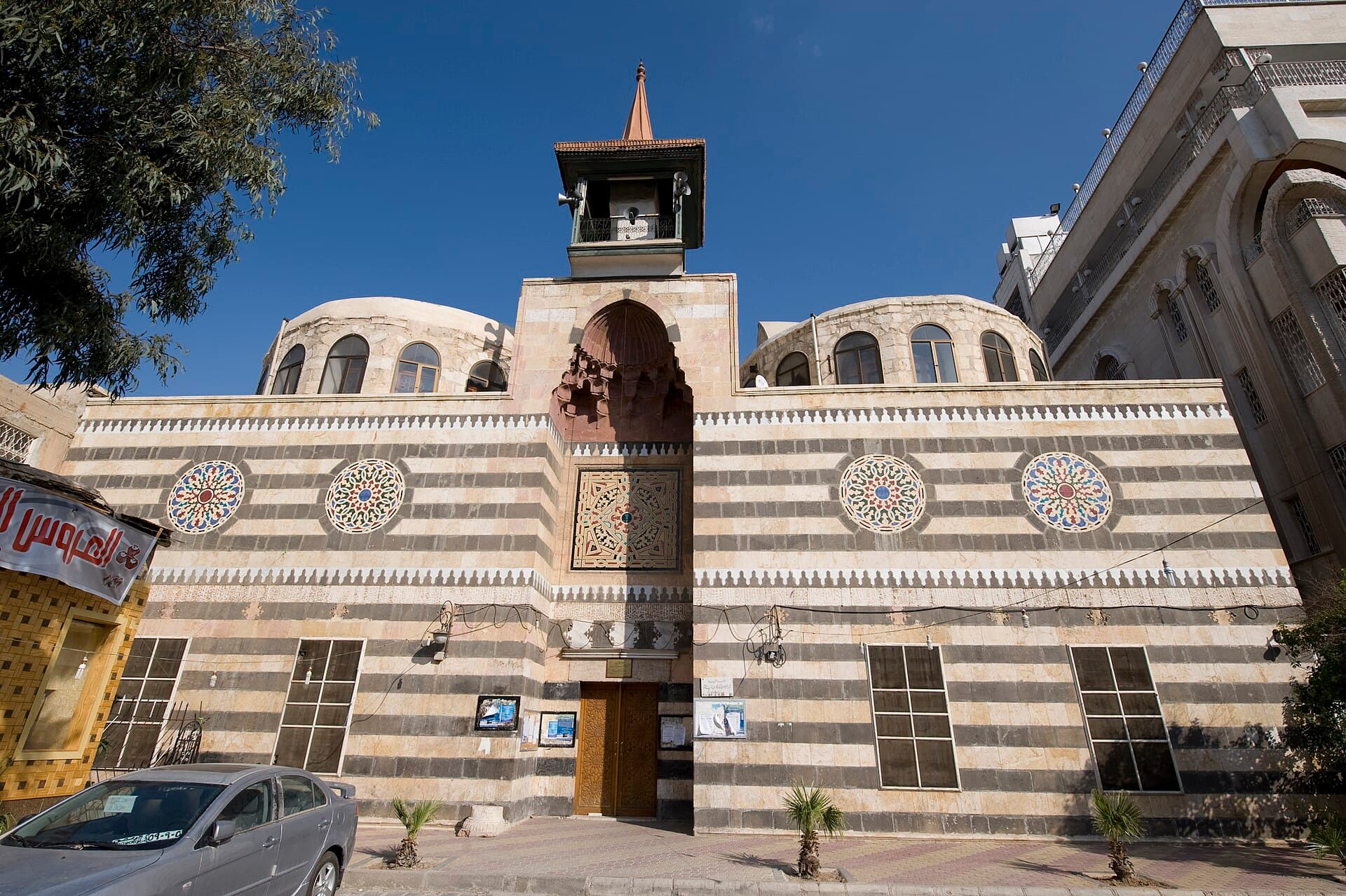 Tanbak Türbesi ve Camii'nin Midan Caddesi'ne bakan onarılmış cephesi: iki kubbe, dört renkli madalyon, kapı üstünde mukarnaslı niş ve ince minare