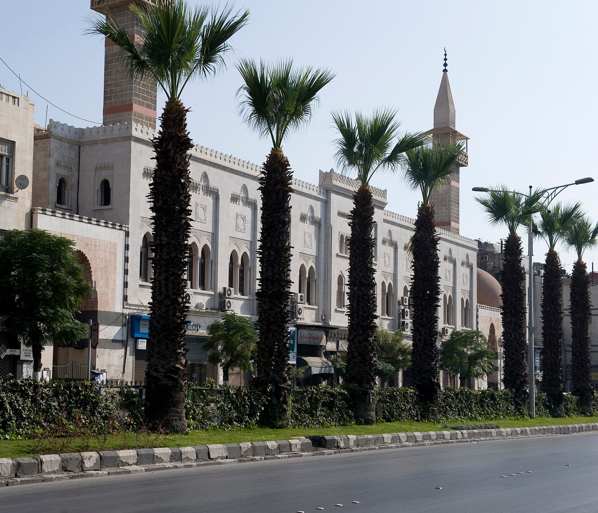 Tankiz Camii'nin Nasır Caddesi'ne bakan beyaz taş cephesi; solda taş-tuğla şeritli eski minare, sağda ince minare ve kubbe, Şam