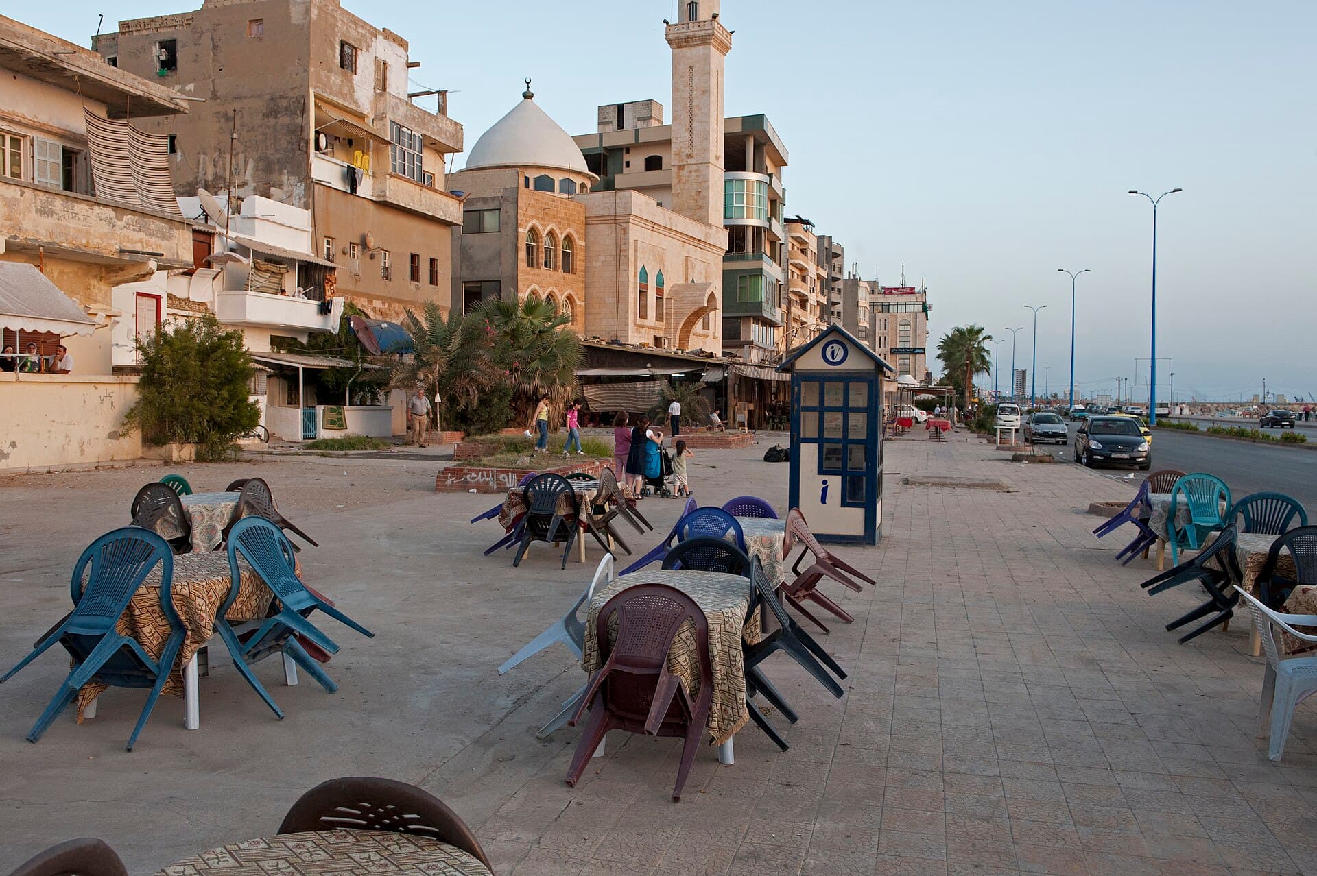 Tartus kornişinde akşamüstü boş kafe masaları, arkada kubbeli ve minareli bir cami ile denize bakan apartmanlar