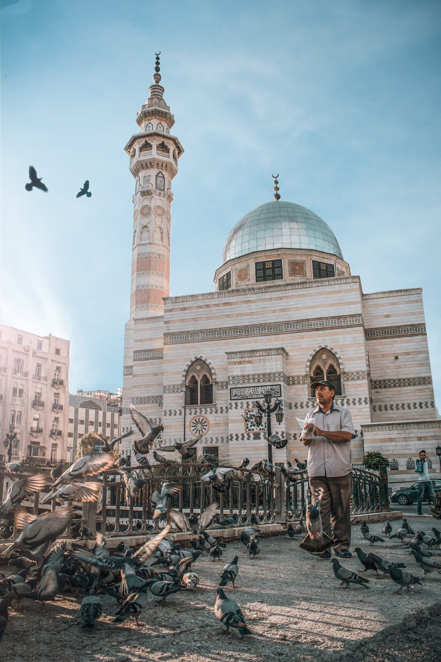 Merce Meydanı'nda Yalbuga Camii: mermer cepheli gövde, madenî kubbe ve uzun minare; önünde güvercin besleyen bir adam