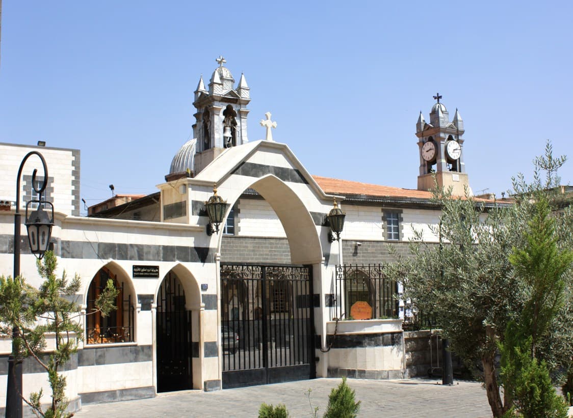 Zeytun Kilisesi'nin siyah-beyaz taş şeritli sivri kemerli taçkapısı; arkada çanlı kule ve saat kulesi, önde zeytin ağaçları