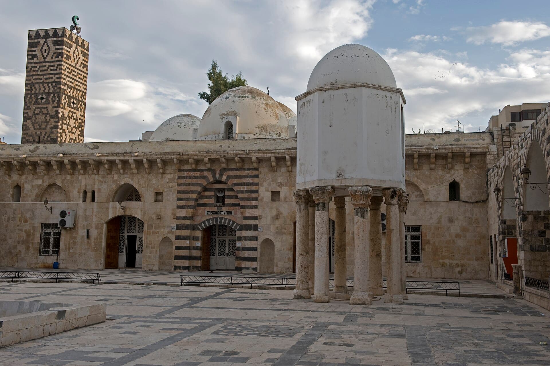 Hama Ulu Camii avlusu: sütunlar üstündeki hazine köşkü, çizgili taçkapı ve desenli kare minare