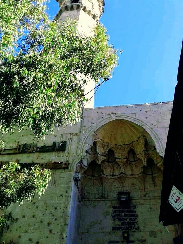 Altun Boğa Camii'nin mukarnaslı taçkapısı ve arkasında yükselen minare; taşta şarapnel izleri (2020)