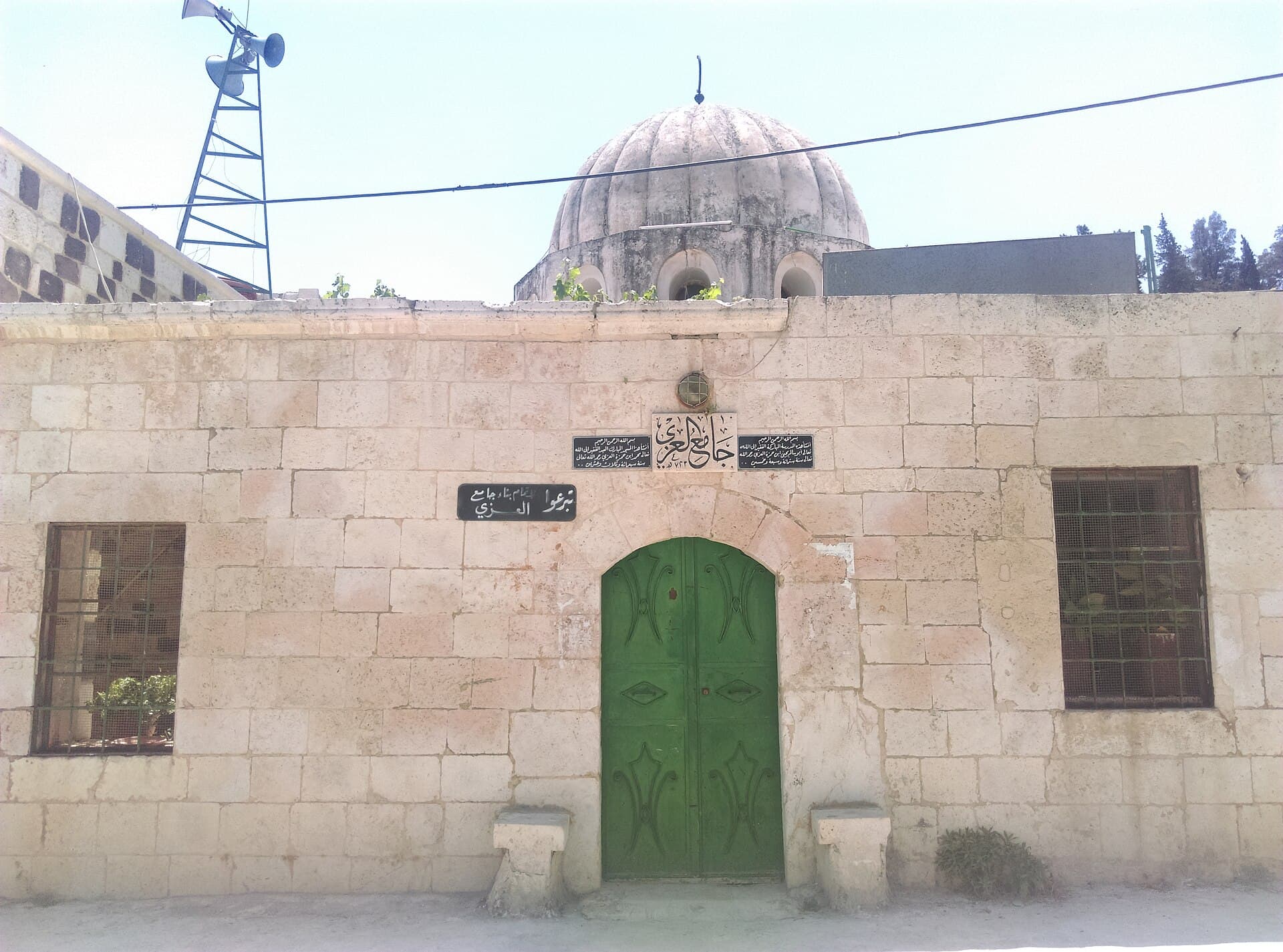 Hama'da Azzi Camii'nin kesme taş cephesi; üstünde caminin adını taşıyan hat levhası bulunan yeşil kapı ve arkada dilimli kubbe