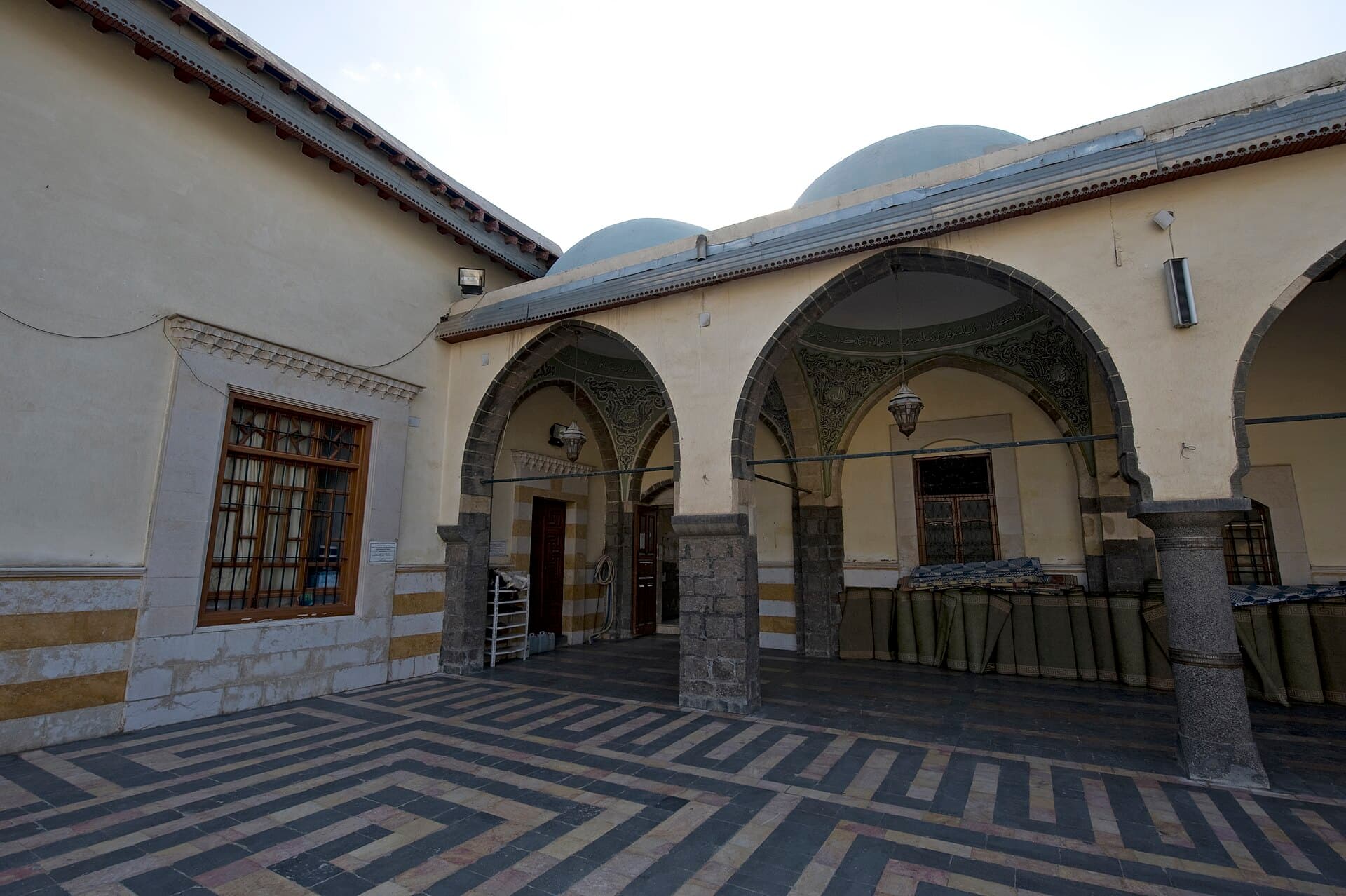 Dakkak Camii'nin avlusu: kubbeli kuzey revağı, kemer üstlerindeki kalem işi ve zikzak desenli taş döşeme, Şam