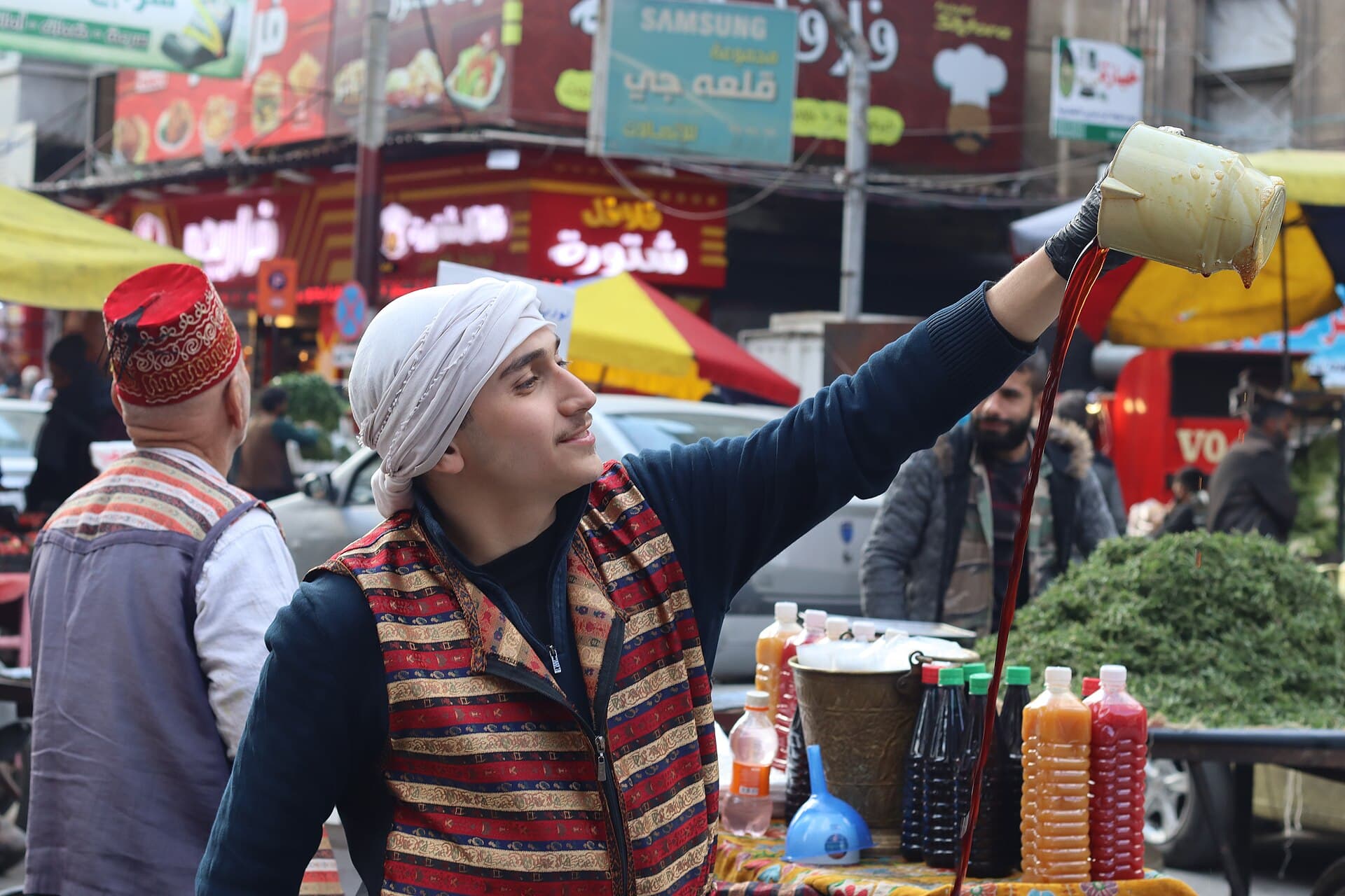 Geleneksel kıyafetli erk sûs satıcısı, çarşıda ibrikten yüksekten döküyor