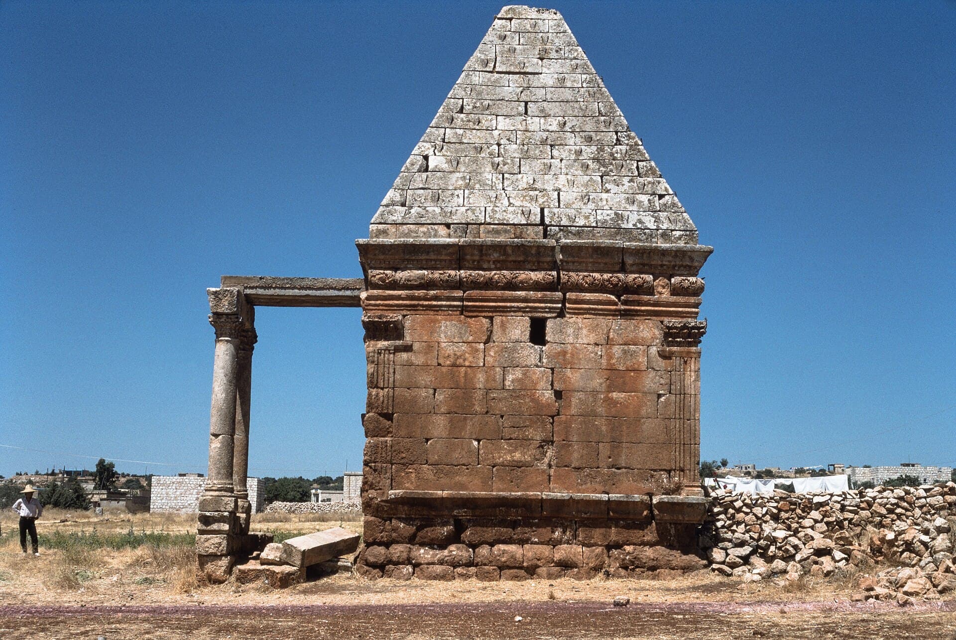 Güney Dana'da piramit çatılı kare taş mezar, köşe plasterleri ve frizi, solda iki sütunun taşıdığı taş kirişli sundurma