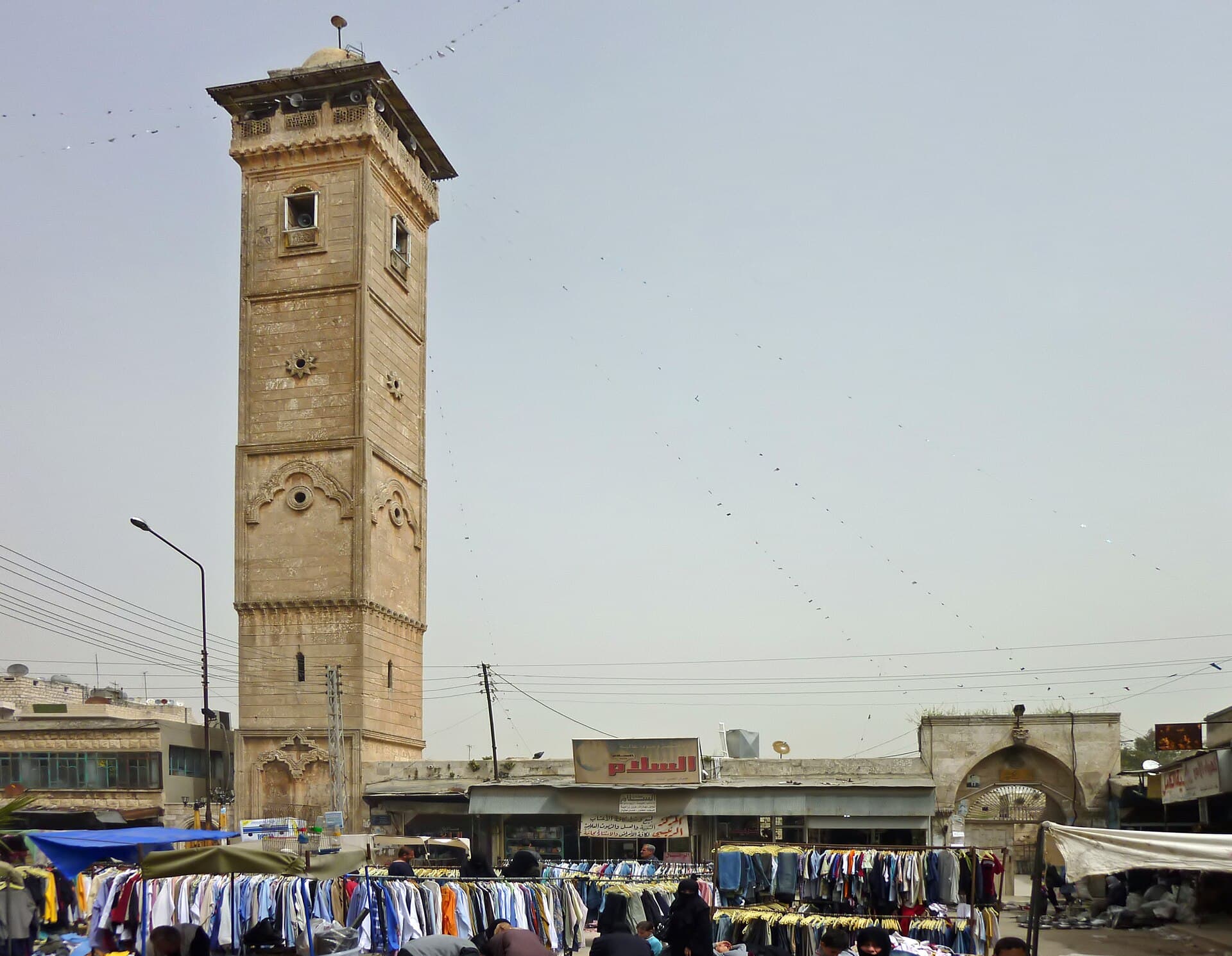 Maarat en-Numan Ulu Camii'nin kare taş minaresi; önünde kasaba pazarının giysi tezgâhları
